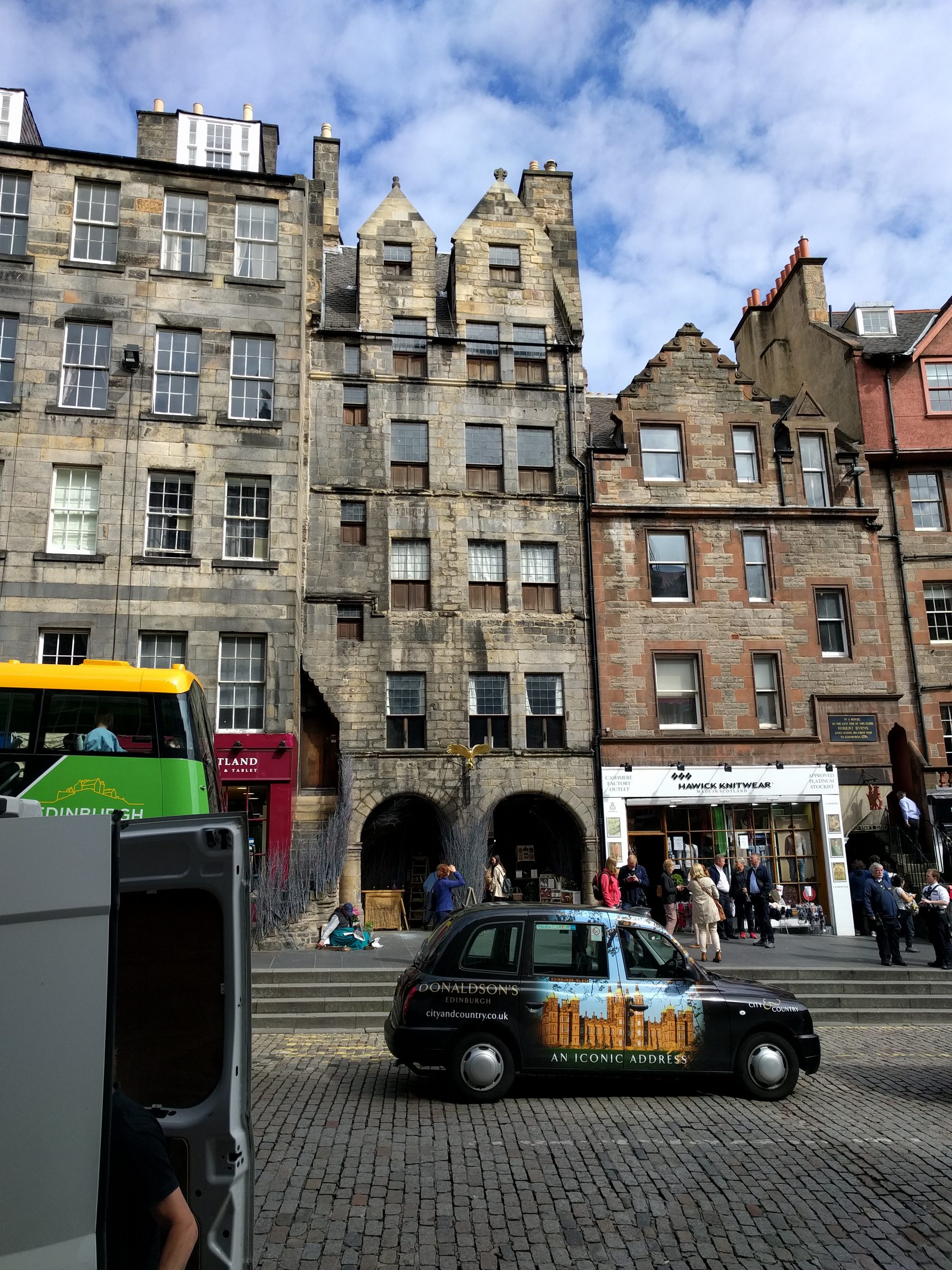 Tall stone tenements and shopfronts along Edinburgh's Royal Mile, a black cab in the foreground