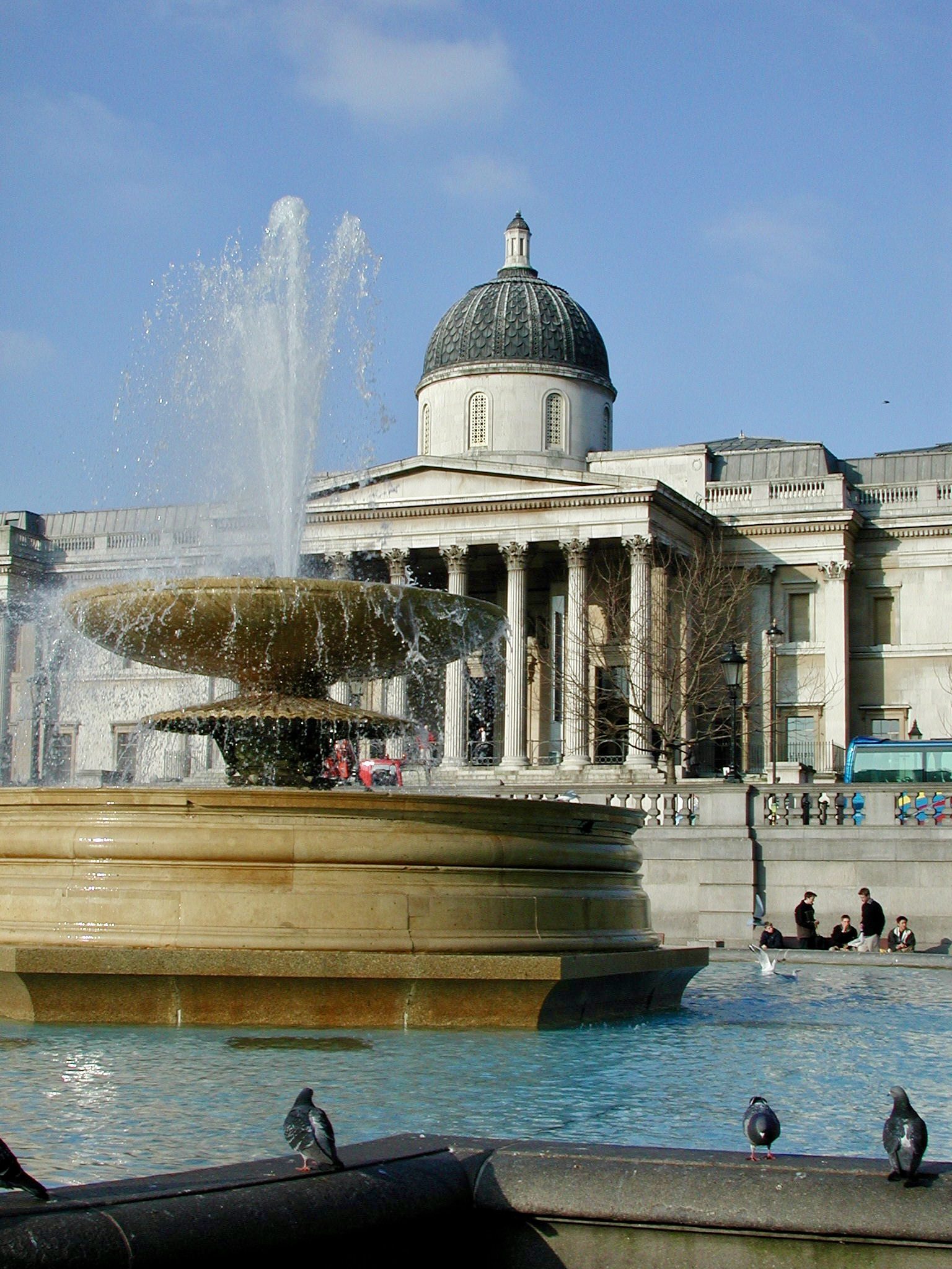A fountain playing in Trafalgar Square with the National Gallery portico behind it