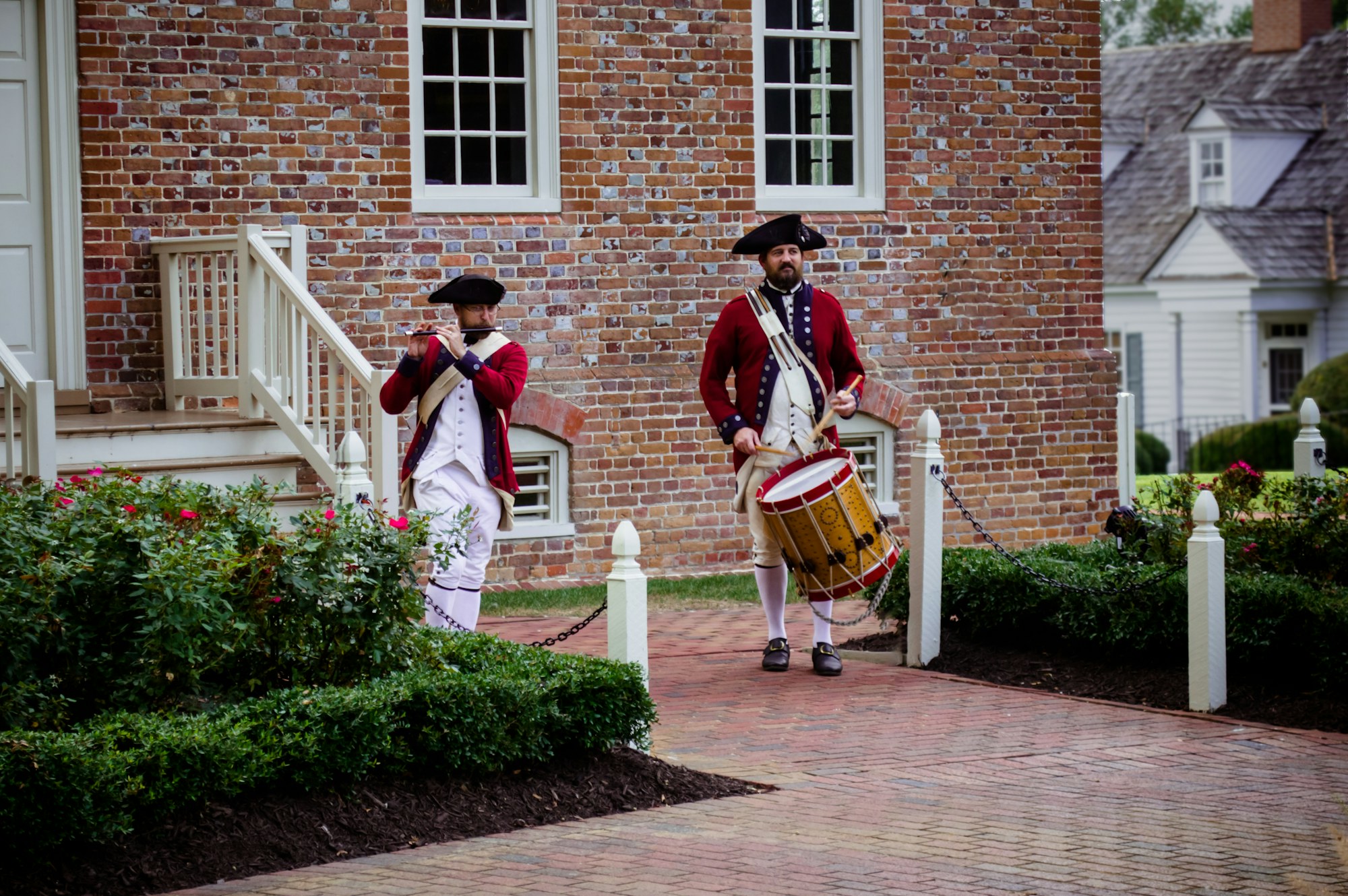 A fifer and a drummer in red coats outside a brick house at Colonial Williamsburg.