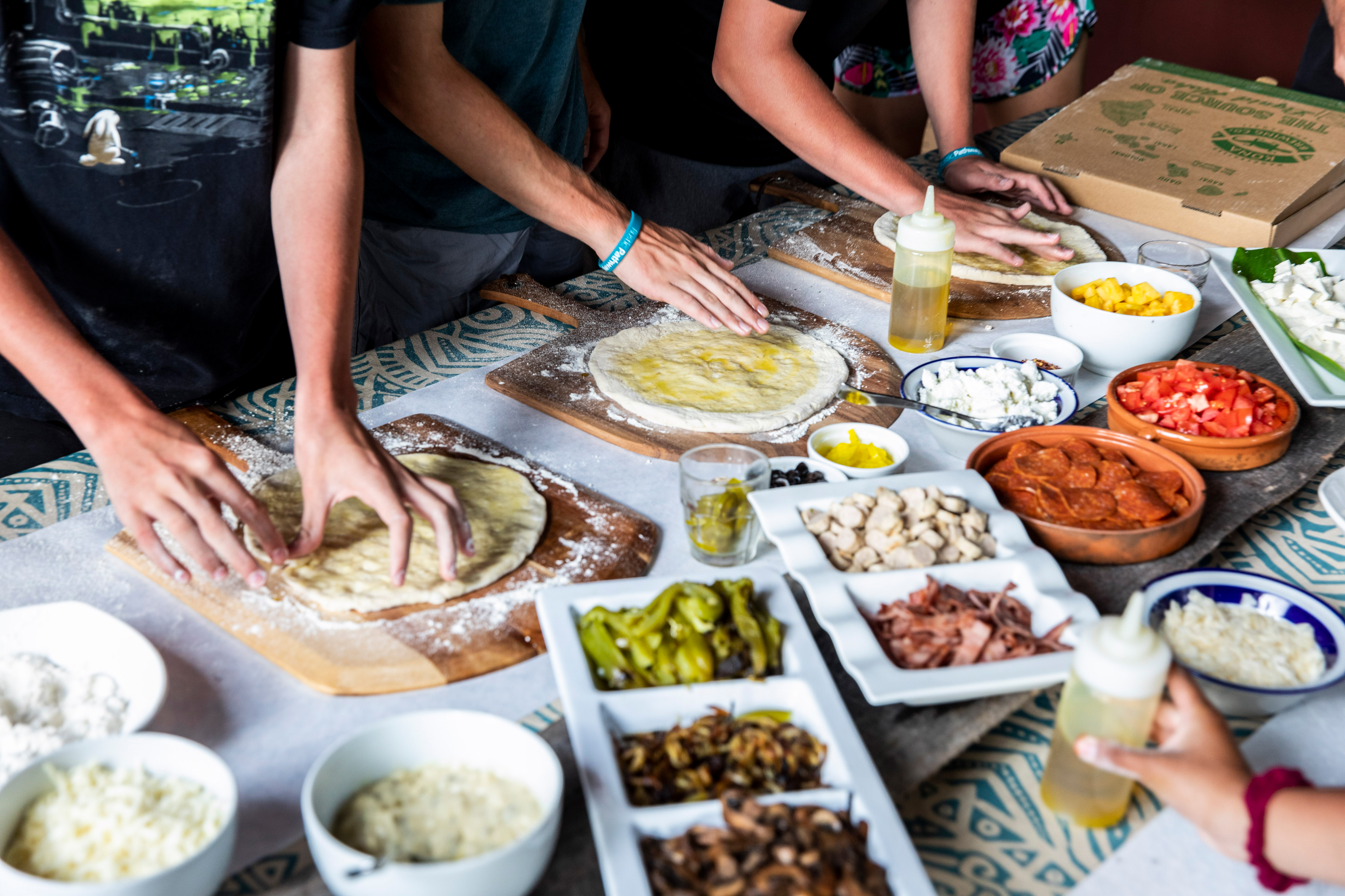Students press out pizza dough on a table topped with bowls of toppings