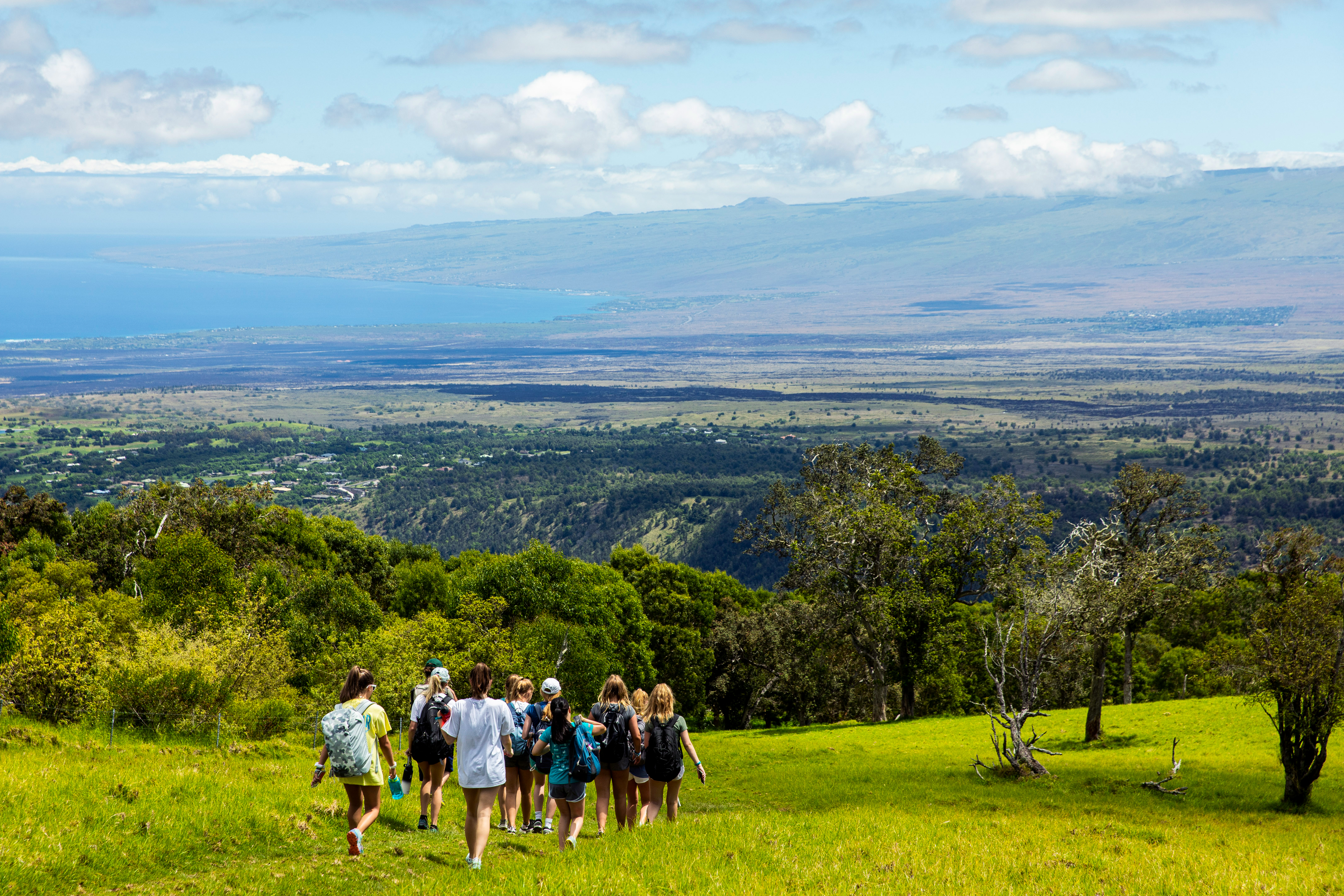 Teen travelers hike down a grassy hillside with ocean views in the distance