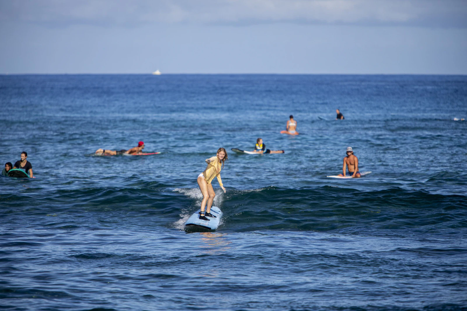 A teen traveler stands up on a surfboard on a small wave while others wait on their boards behind