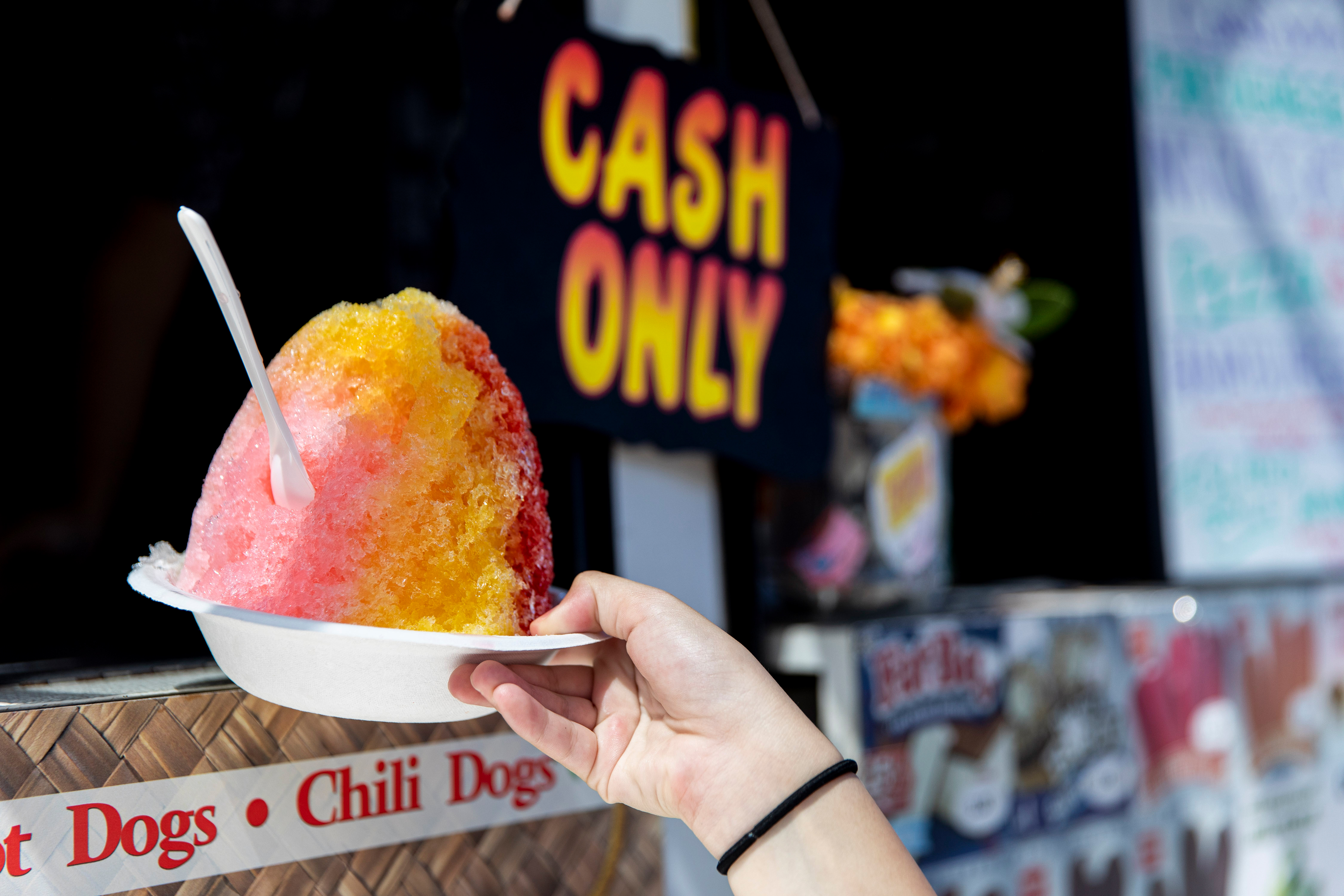 A hand holds a shave ice in front of a "Cash Only" sign at a food stand