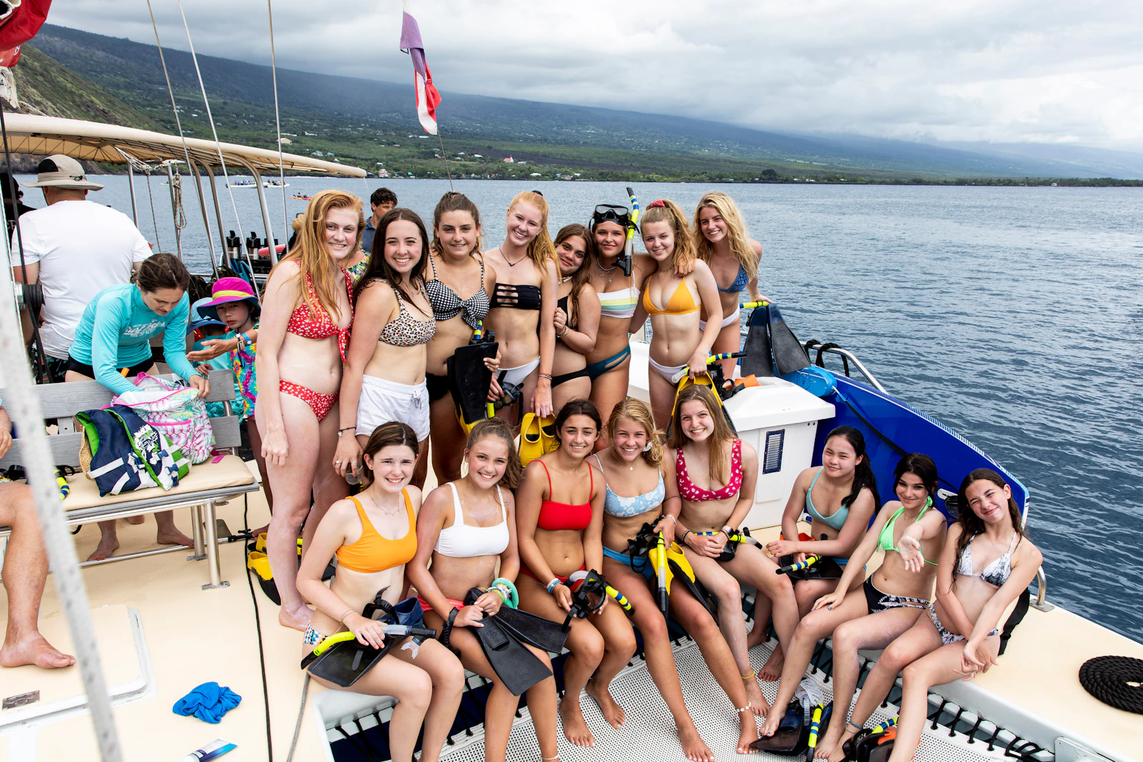 Teen travelers in swimsuits pose with fins and snorkels on the deck of a boat, green mountains behind