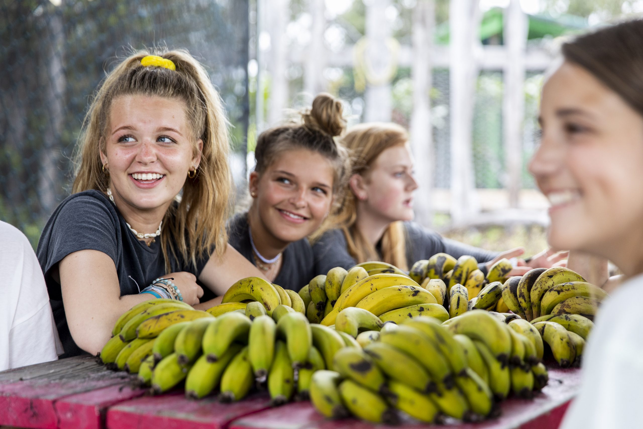 Photo of girls sitting on a bench with bananas on the table in the center.
