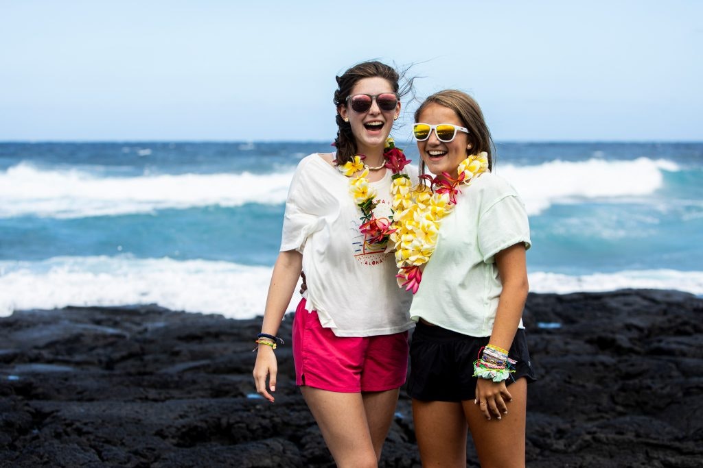 Two girls are standing in front of the ocean. They are wearing sunglasses, white T-Shirts and Lei.