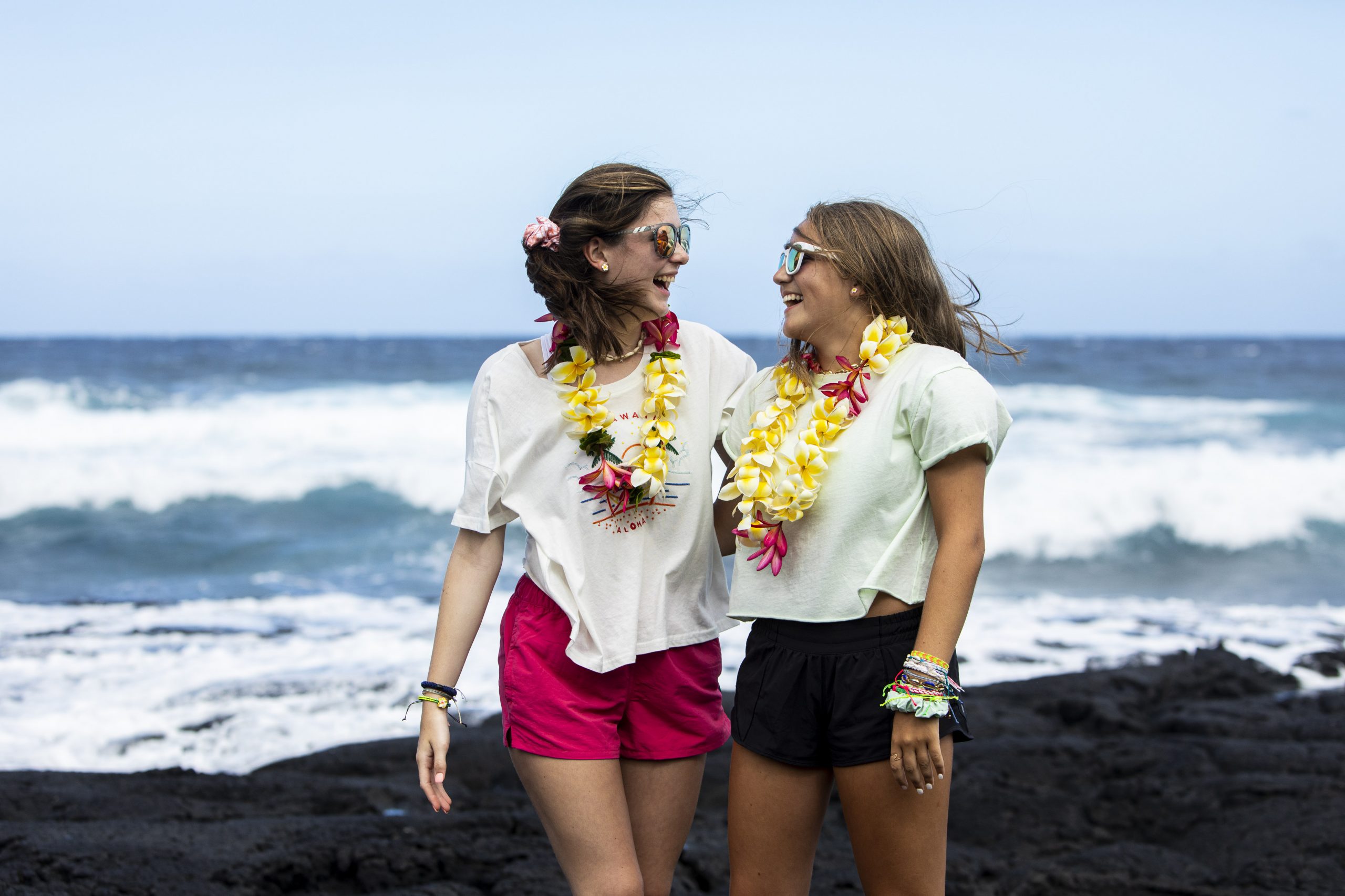 Teen travelers wearing flower leis laugh together on a black sand beach