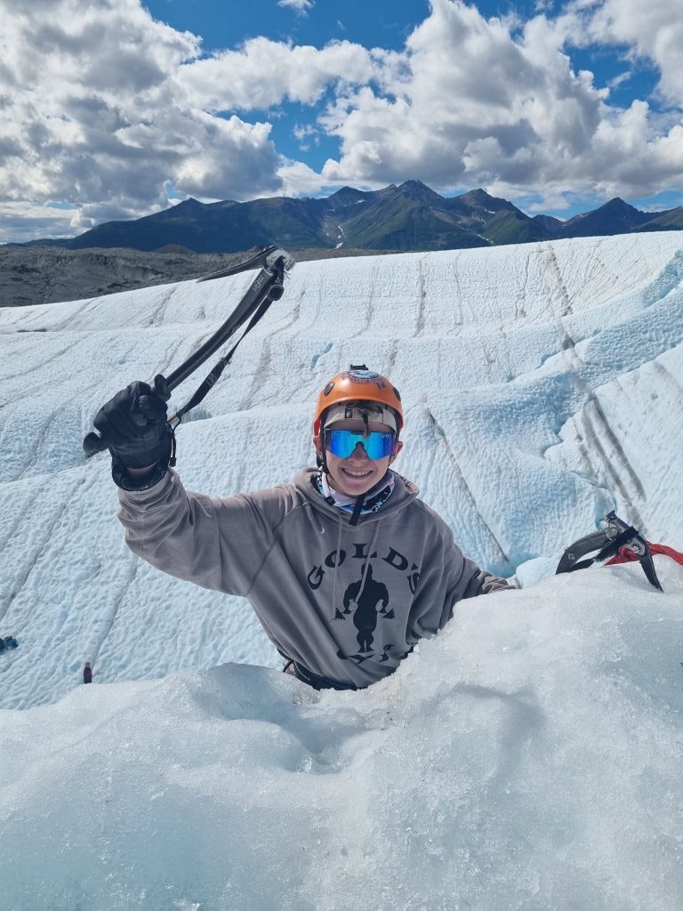 A student wearing a helmet and goggles, and carrying an icepick smiles at the camera while ice climbing during a teen tour in the USA.