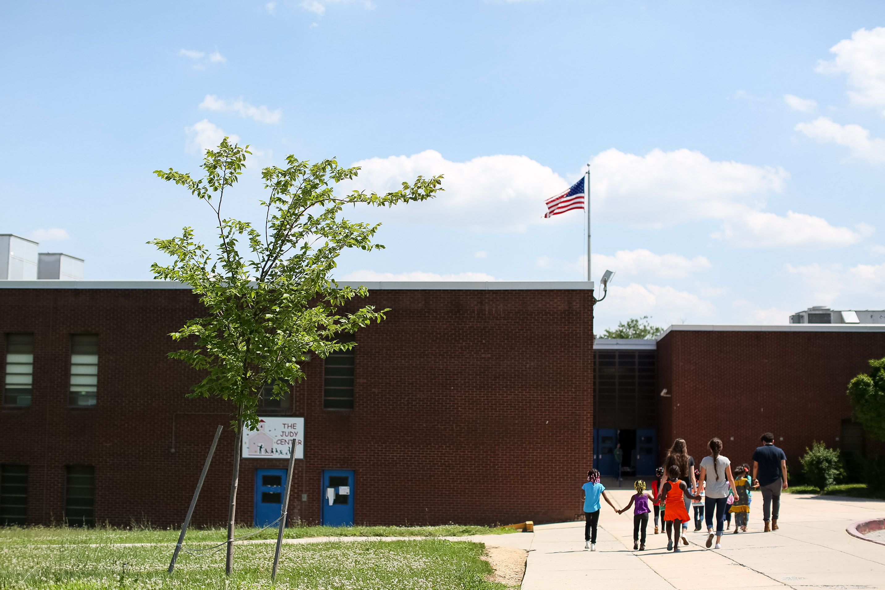 Families walk hand in hand toward a brick school building under an American flag