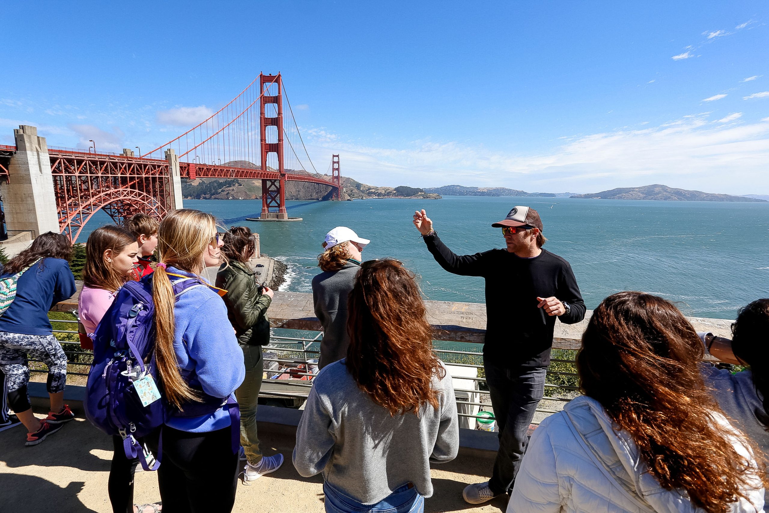 A guide gestures toward the Golden Gate Bridge as teens listen at a viewpoint