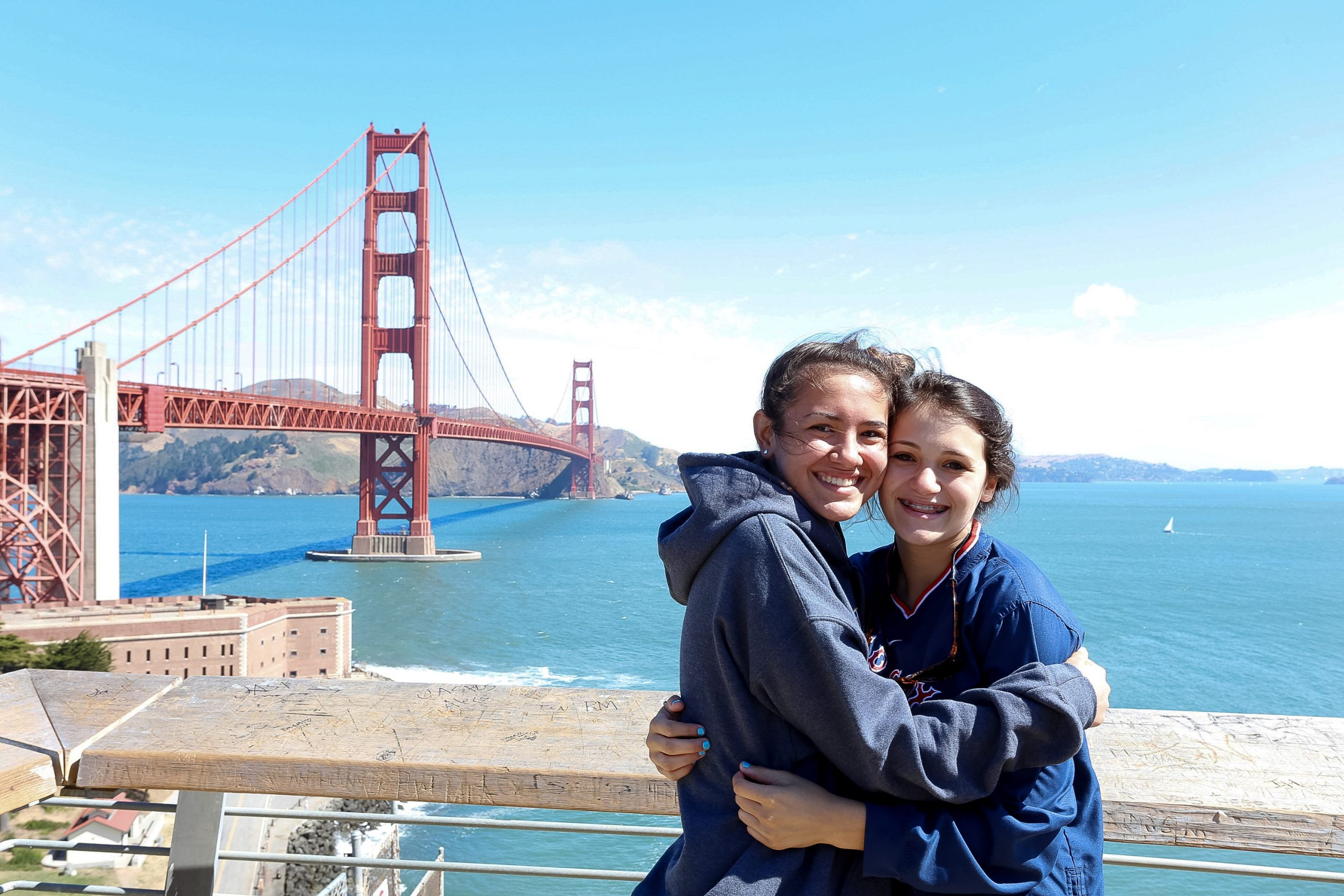 Two friends smiling in front of the Golden Gate Bridge in San Francisco on a clear day.