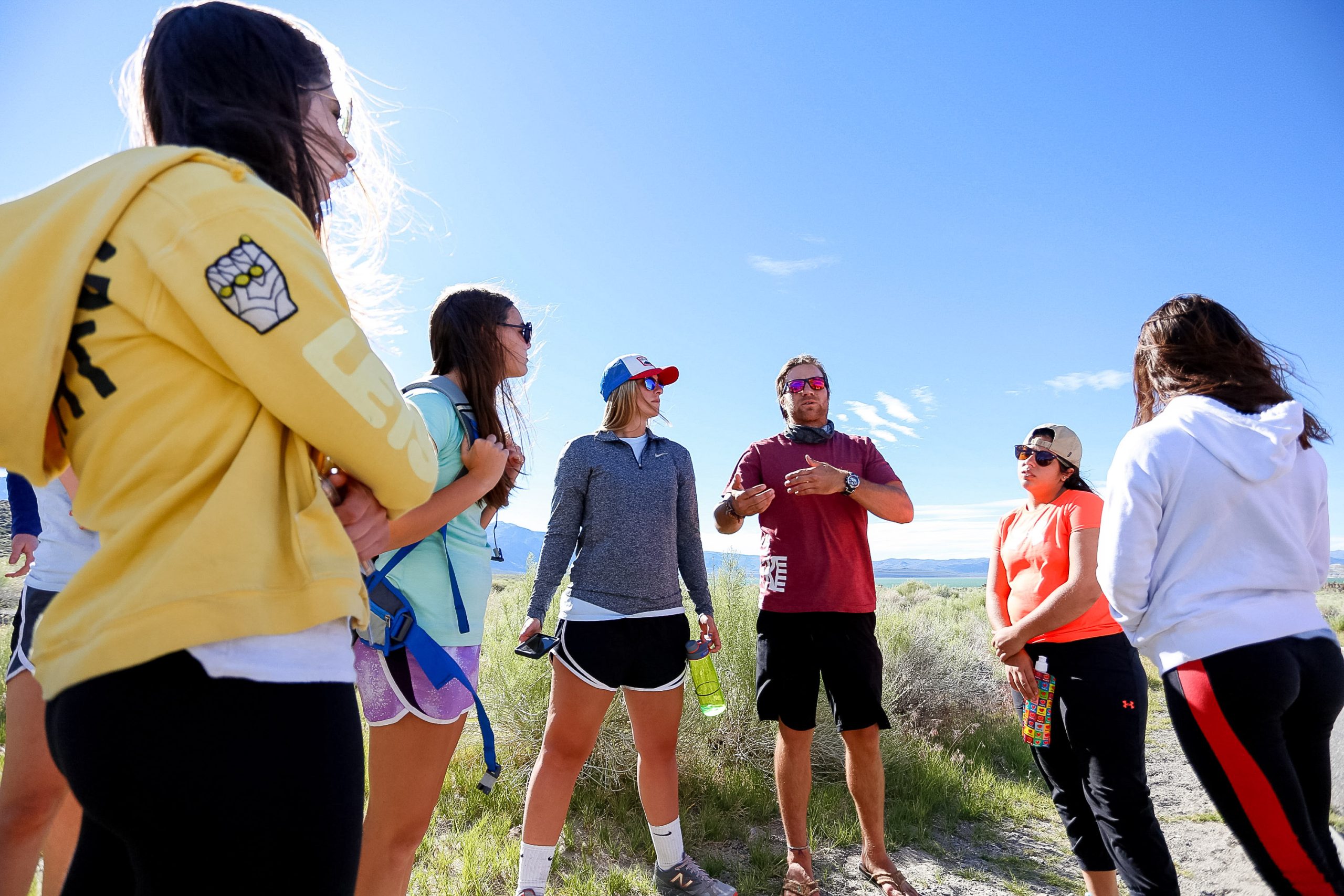 Students touring a university in the United States gather outdoors listening to a guide on a grassy hillside