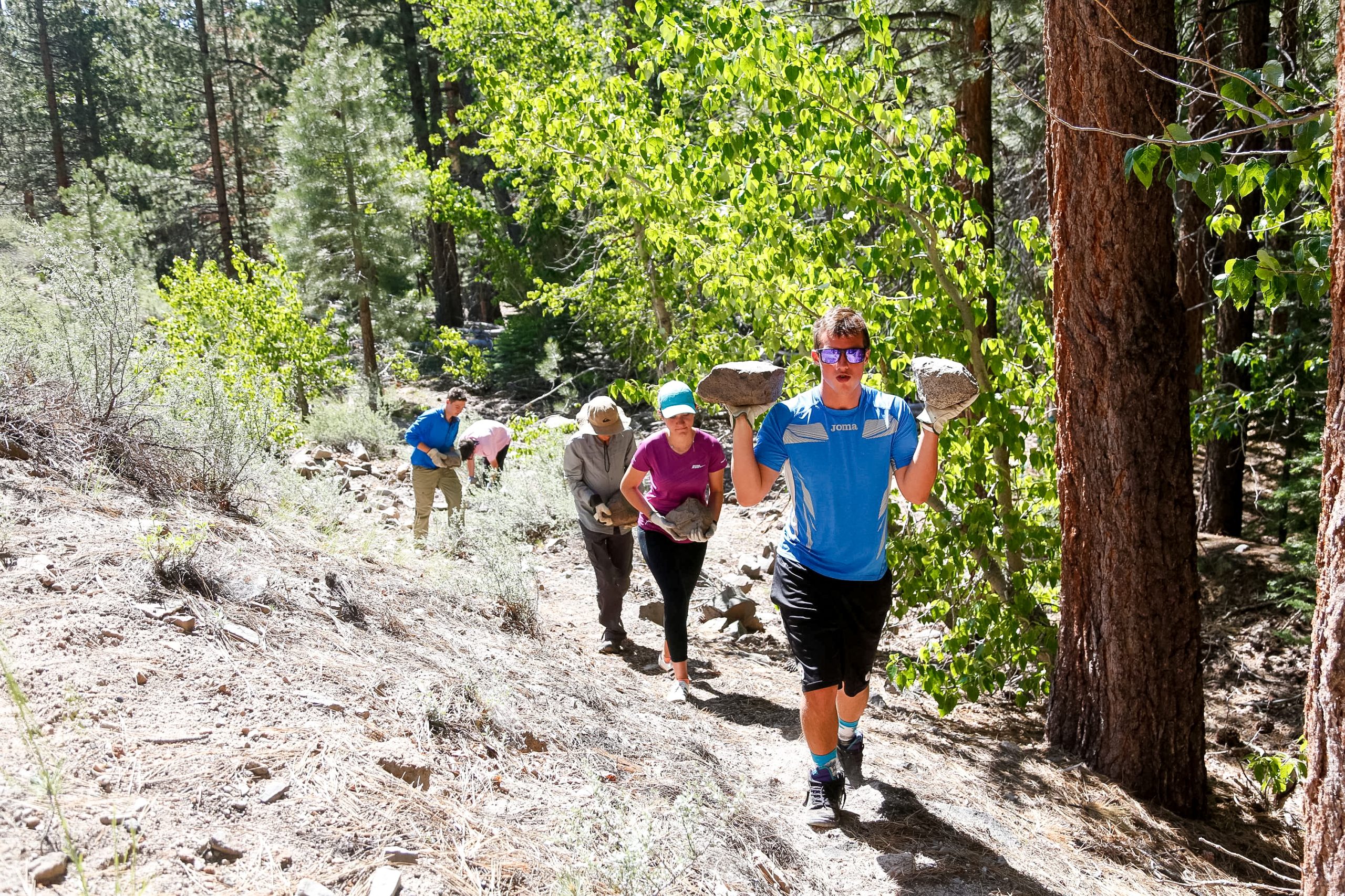 A group of hikers walking up a trail, with one person carrying large rocks on their shoulders.