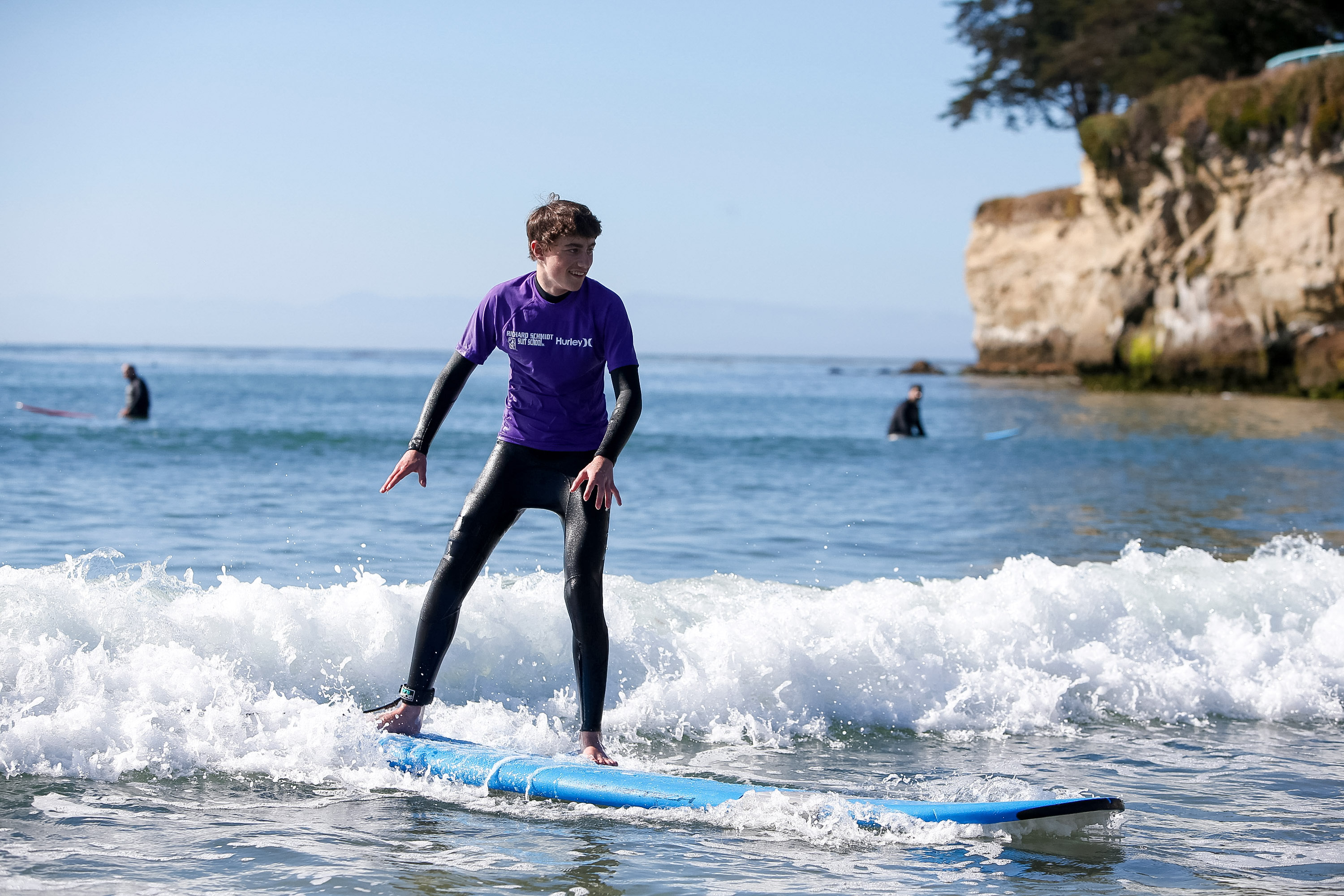 Students ride small waves while learning to surf near a rocky coastline