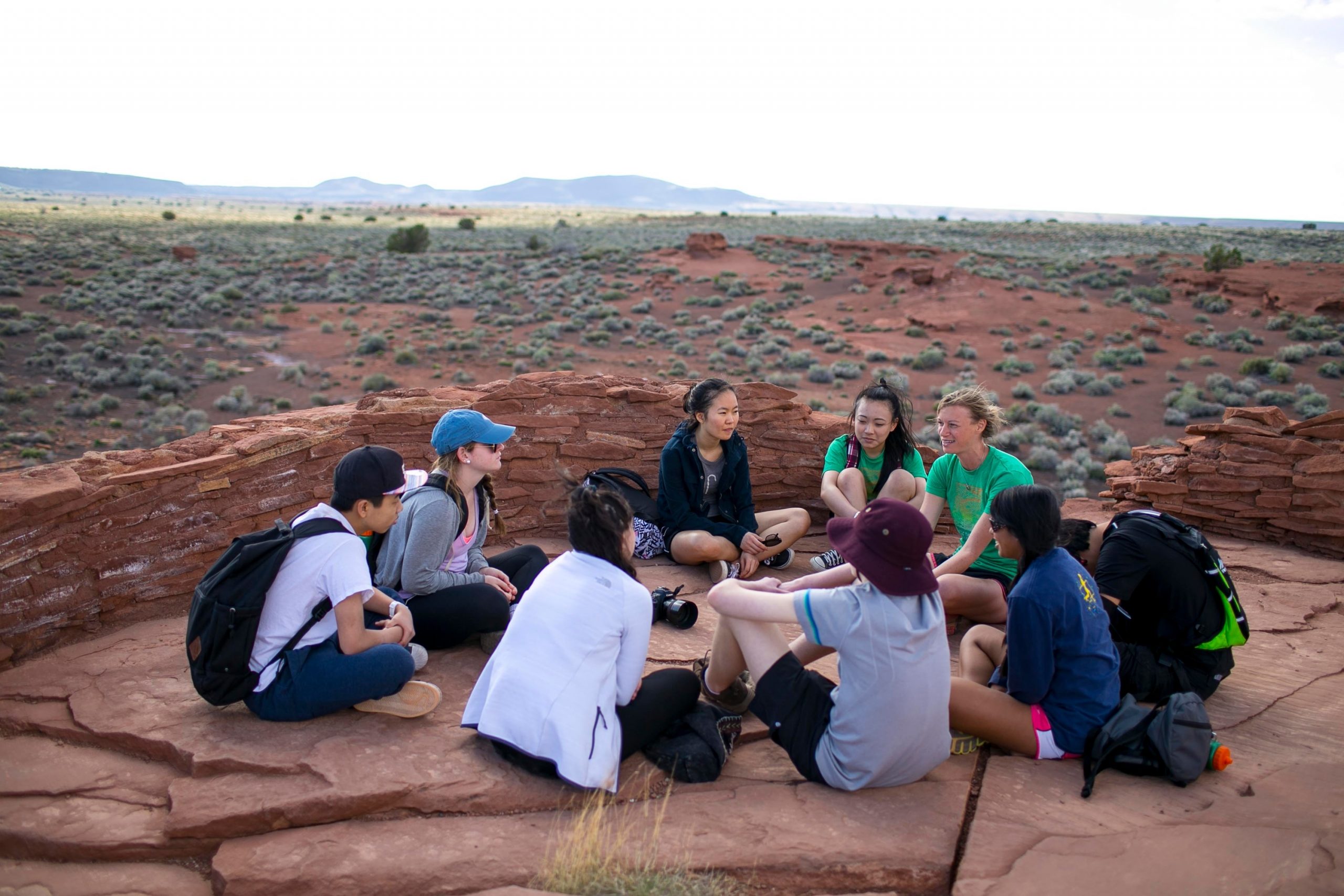 Students sit in a circle atop red rock overlooking a desert landscape
