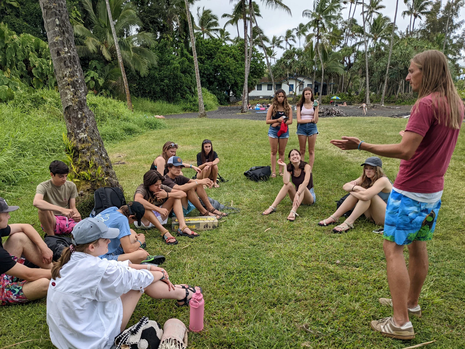 An adult talks to teen travelers sitting on the grass under tall palm trees