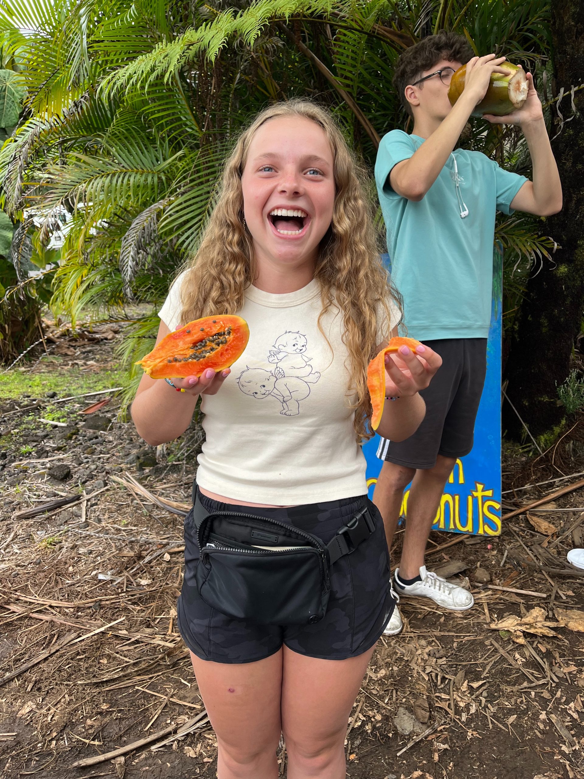 A teen traveler laughs holding two halves of a papaya while a friend drinks from a coconut behind her