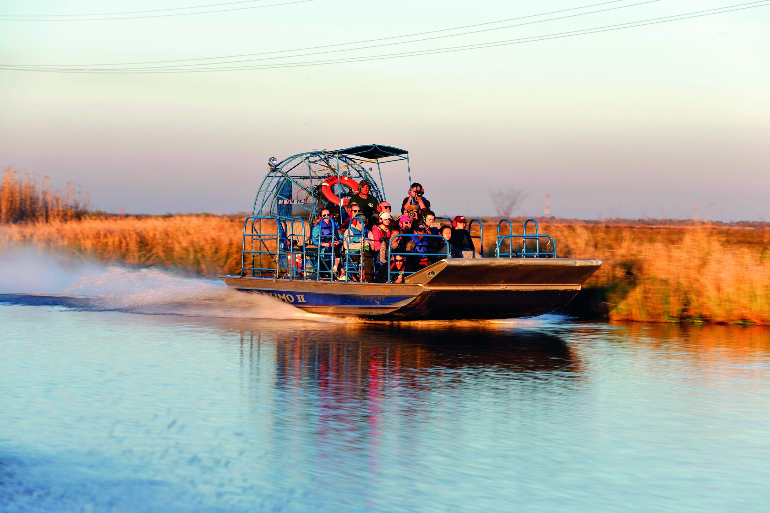 A group of people, wearing life jackets, take an adventurous airboat ride through a marshy wetland, with golden grasses and still water reflecting the sky.