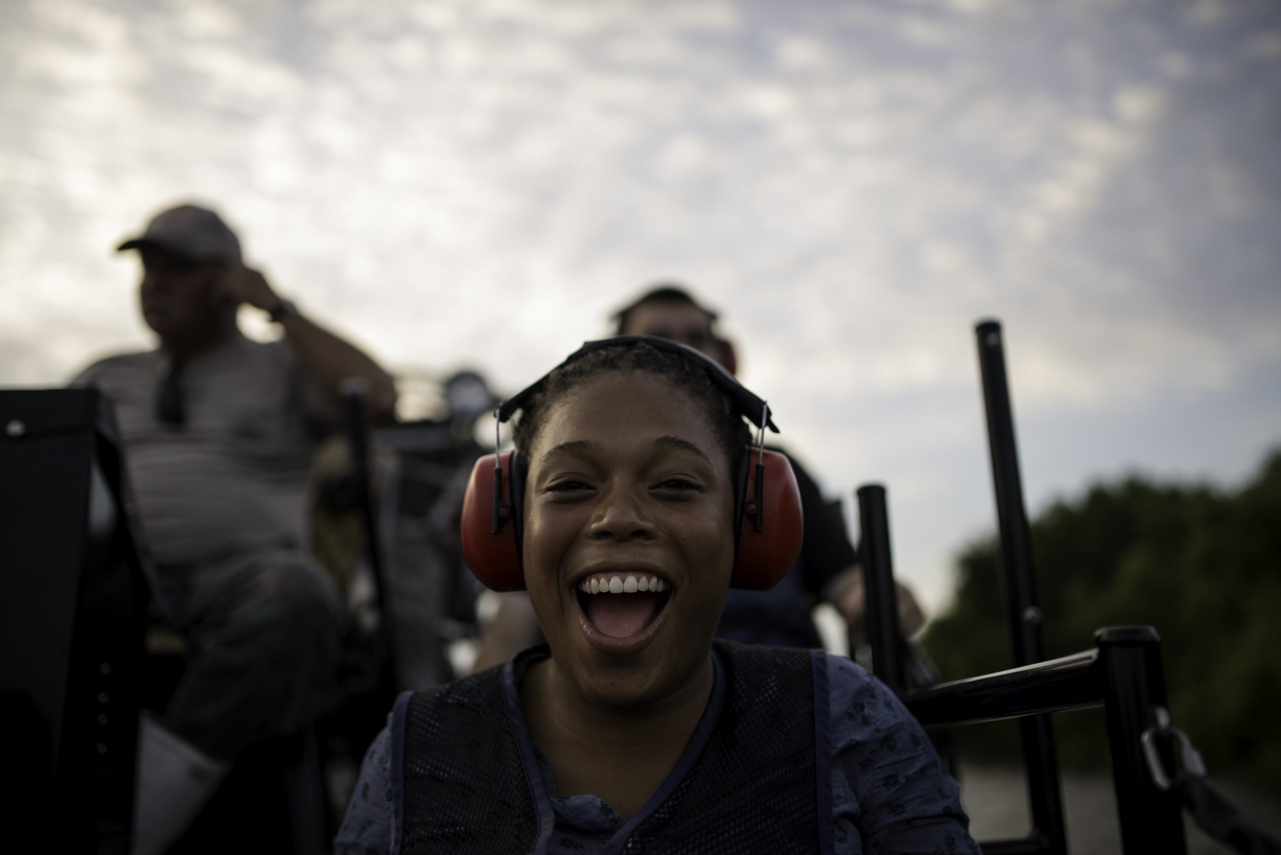 fun facts about new orleans. A young person wearing red ear protection smiles widely while riding on a vehicle, with others seated behind them. The background shows a cloudy sky and blurred greenery, suggesting an outdoor adventure.