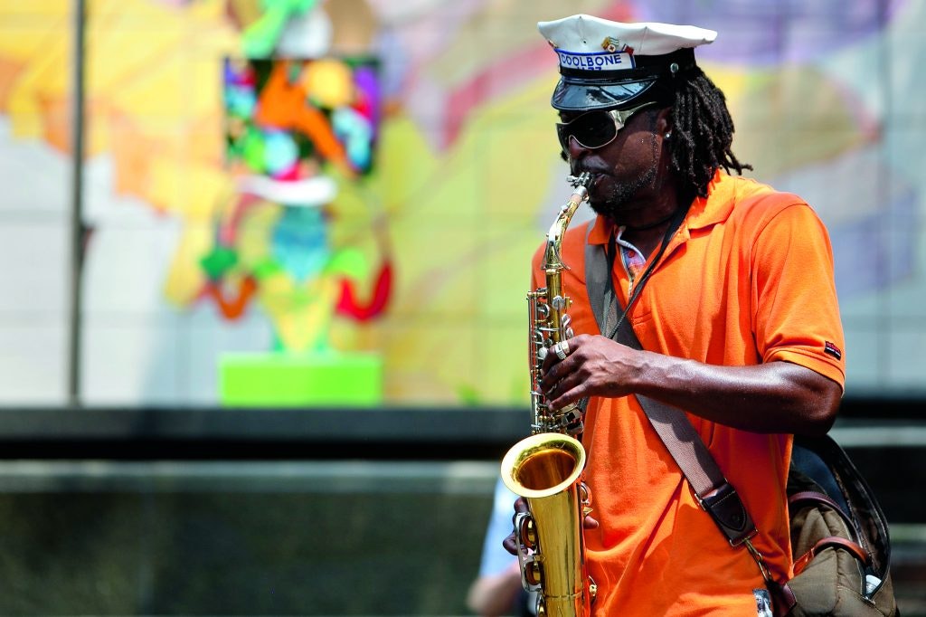 A Black musician wearing sunglasses and a white sailor-style hat labeled "COOLBONE" plays the saxophone in front of a colorful mural, embodying the rich musical heritage of New Orleans.