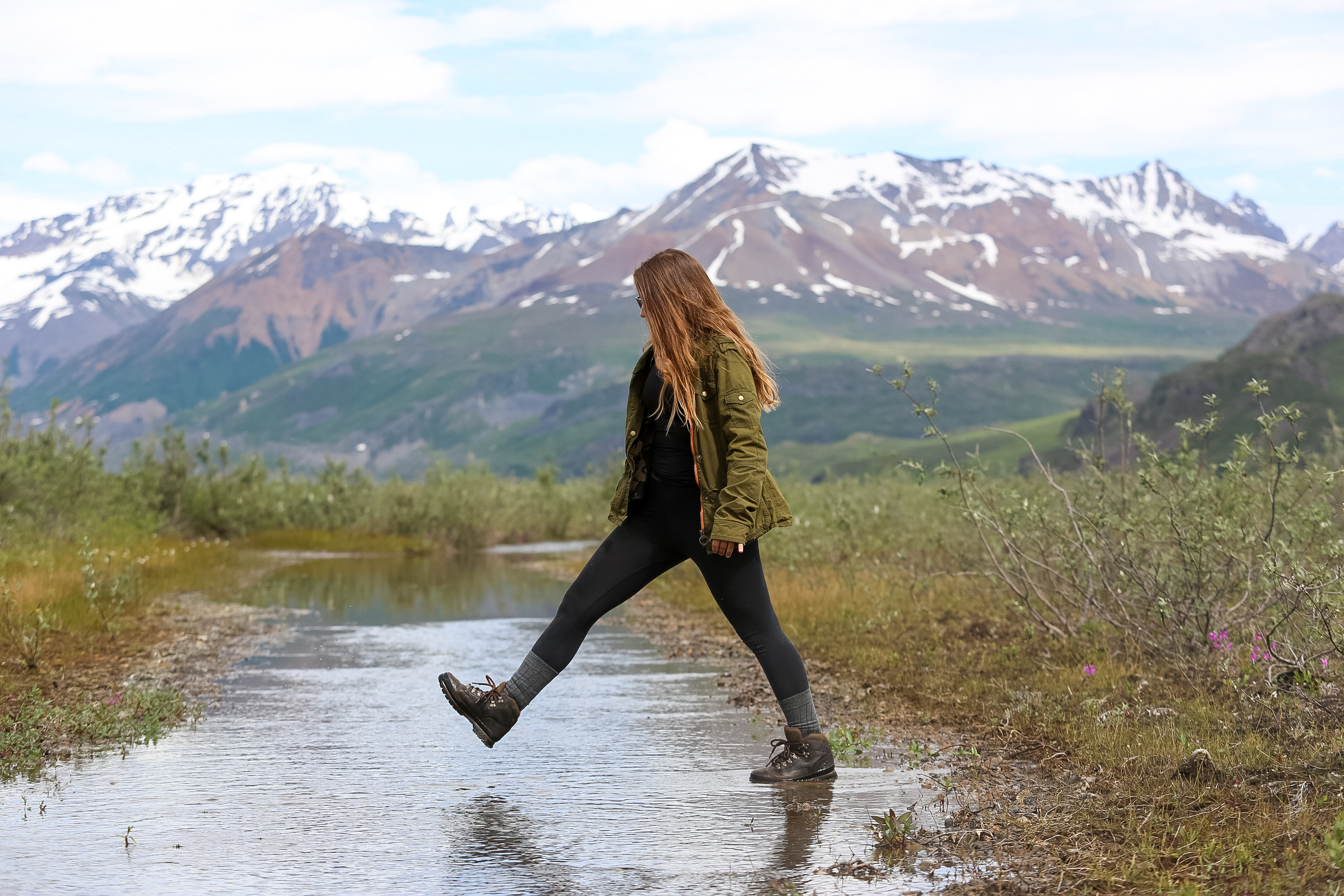 A student steps across a shallow stream with snow-capped mountains behind