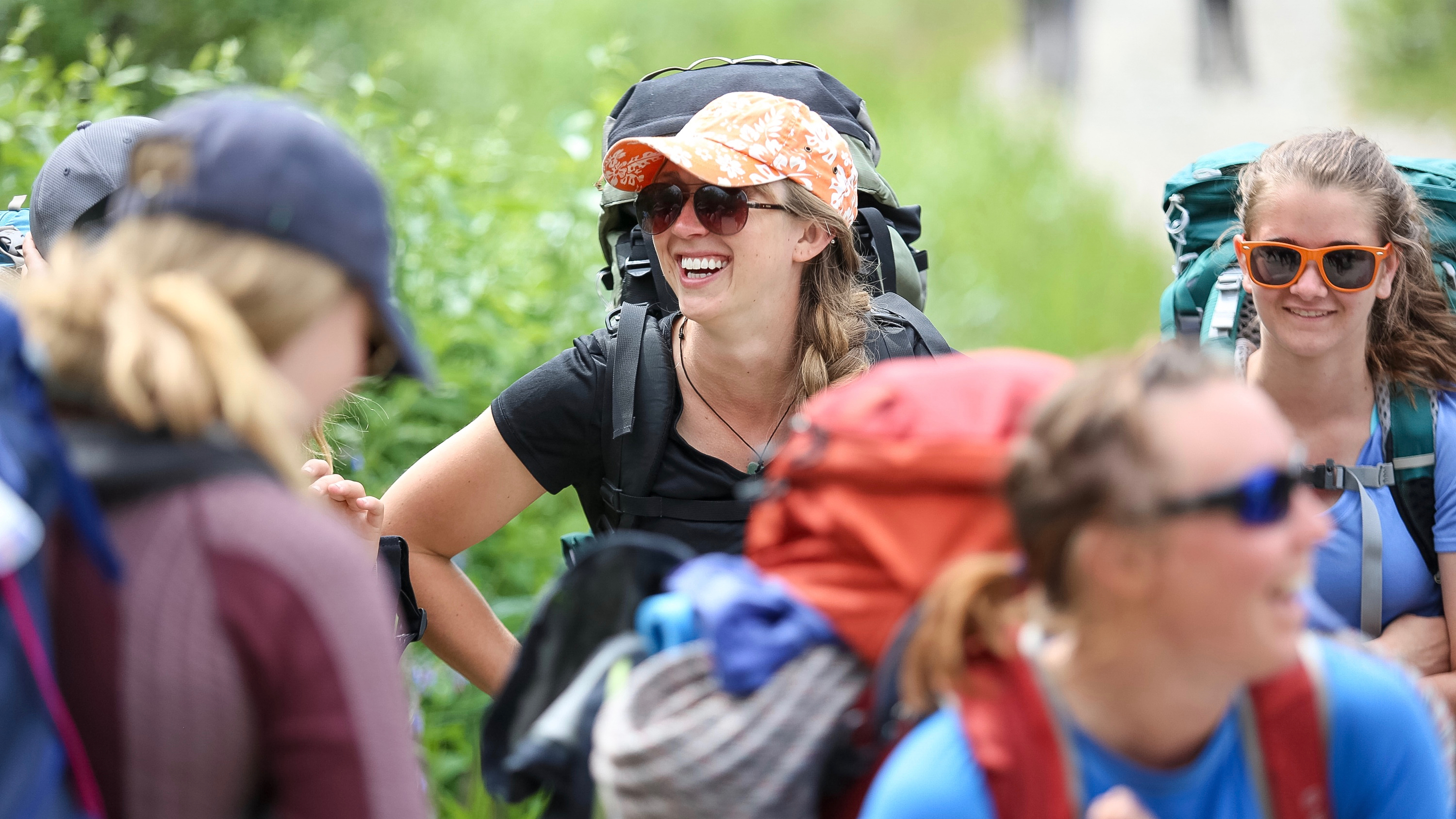Students wearing backpacking packs laugh together on a hike through green brush