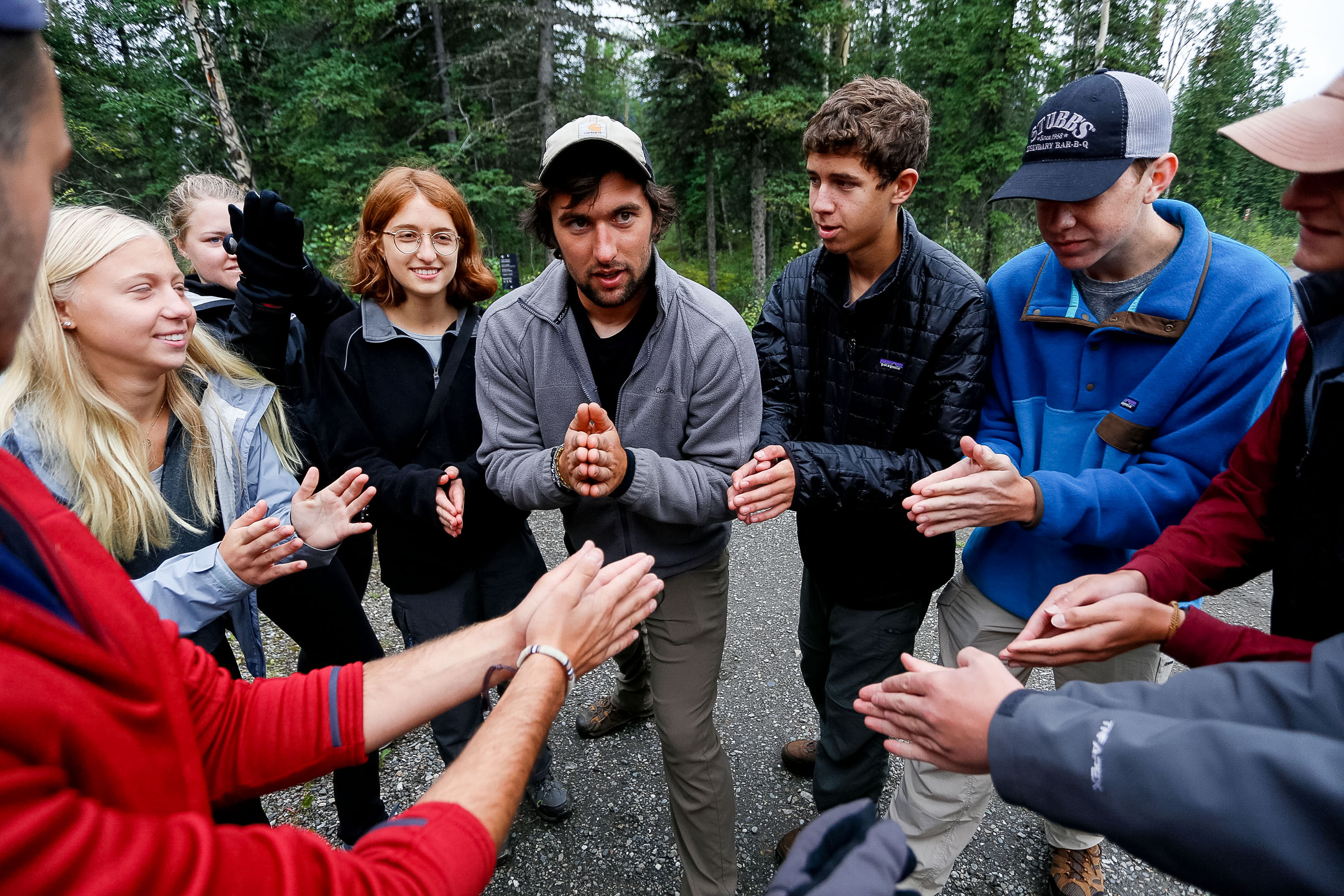Students and a leader stand in a circle on a gravel road in a forest in the United States, clasping hands