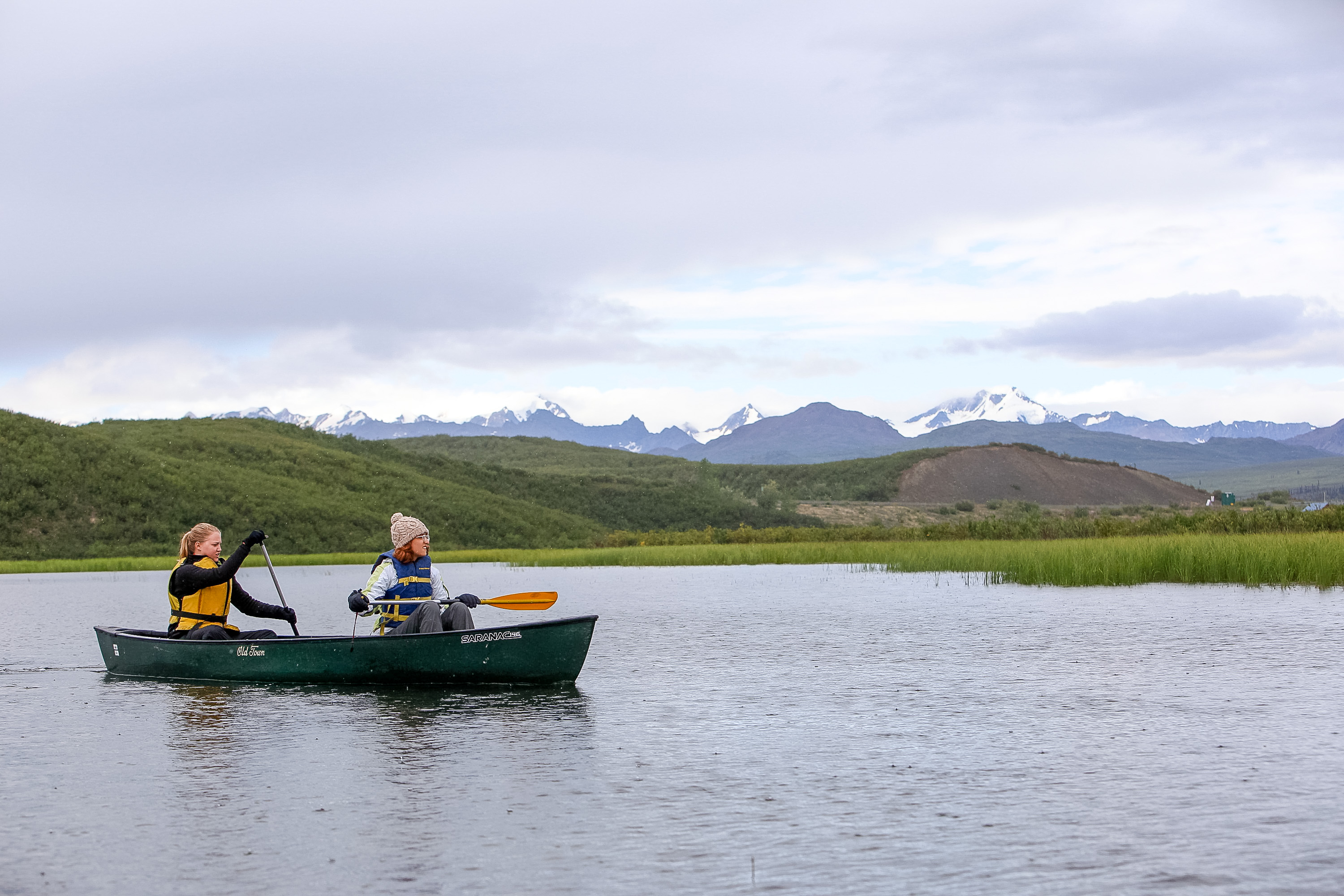 Students paddle a canoe across a still lake with snow-capped mountains behind