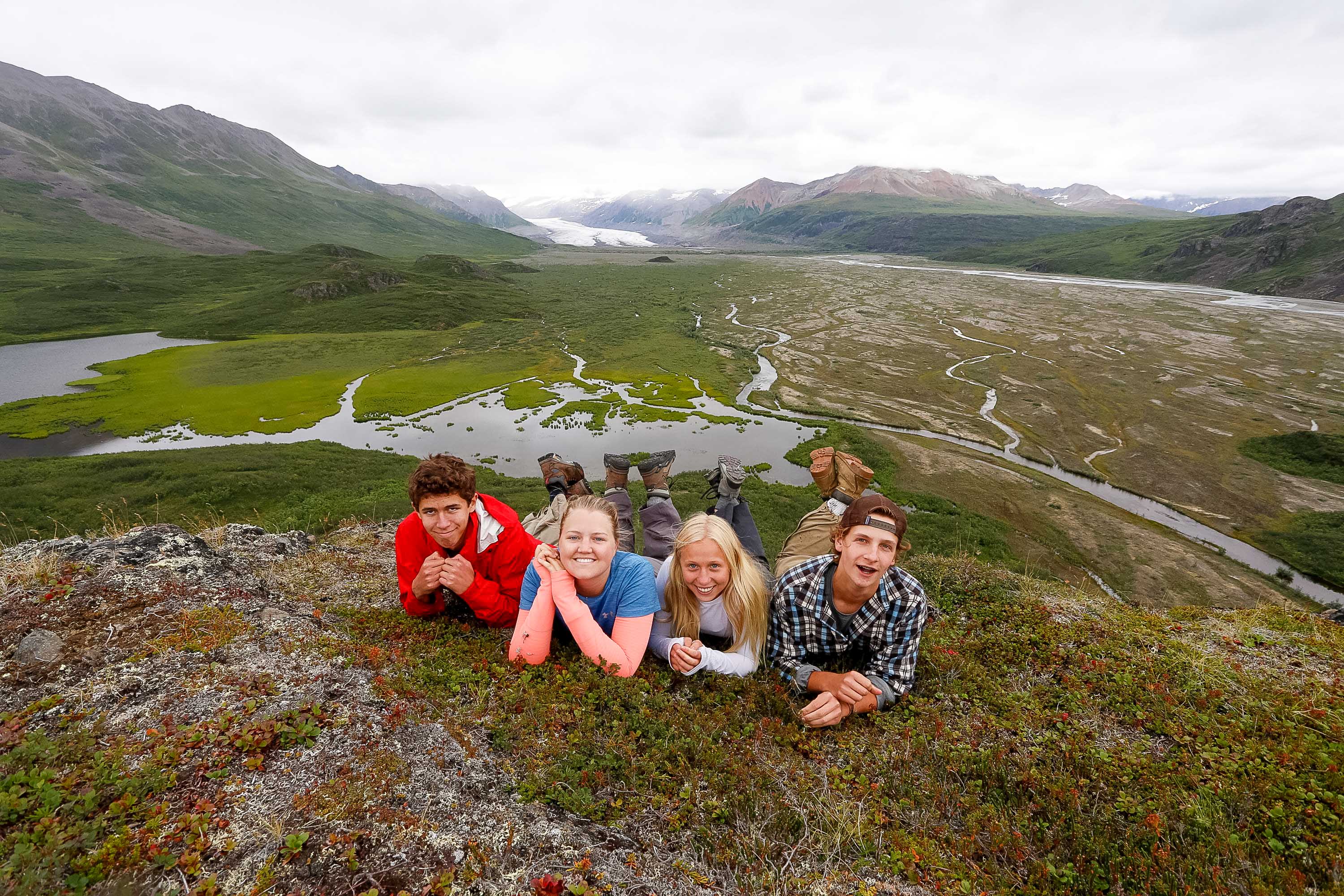 Students lie on a grassy ridge overlooking a braided river valley and mountains