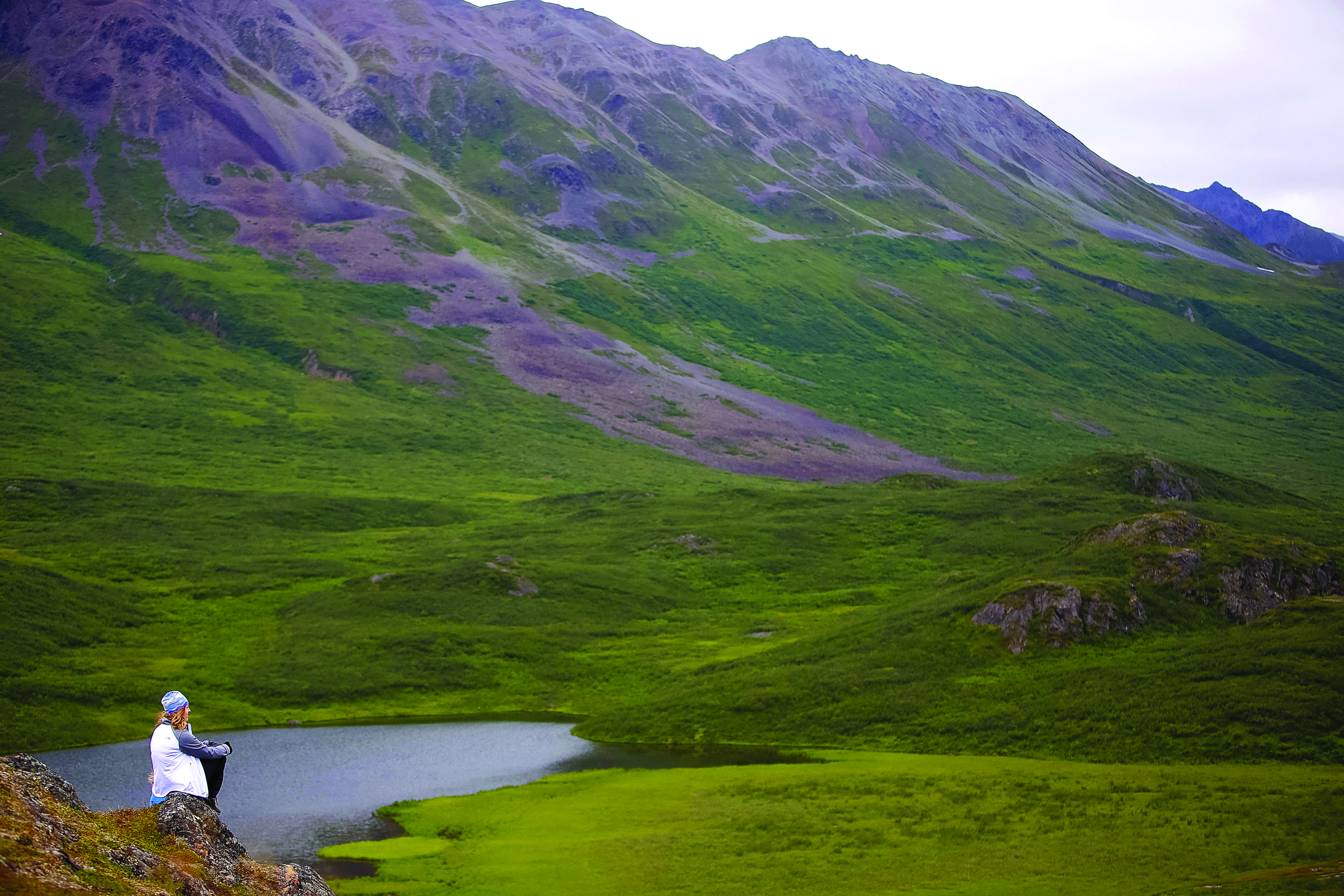 A student sits on a rocky outcrop overlooking a mountain lake in Alaska