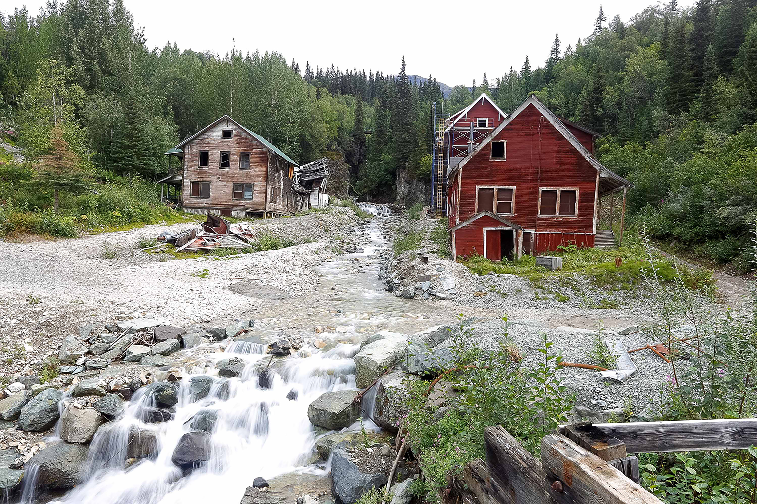 Weathered wooden mining buildings stand along a rocky stream in a forested Alaskan valley