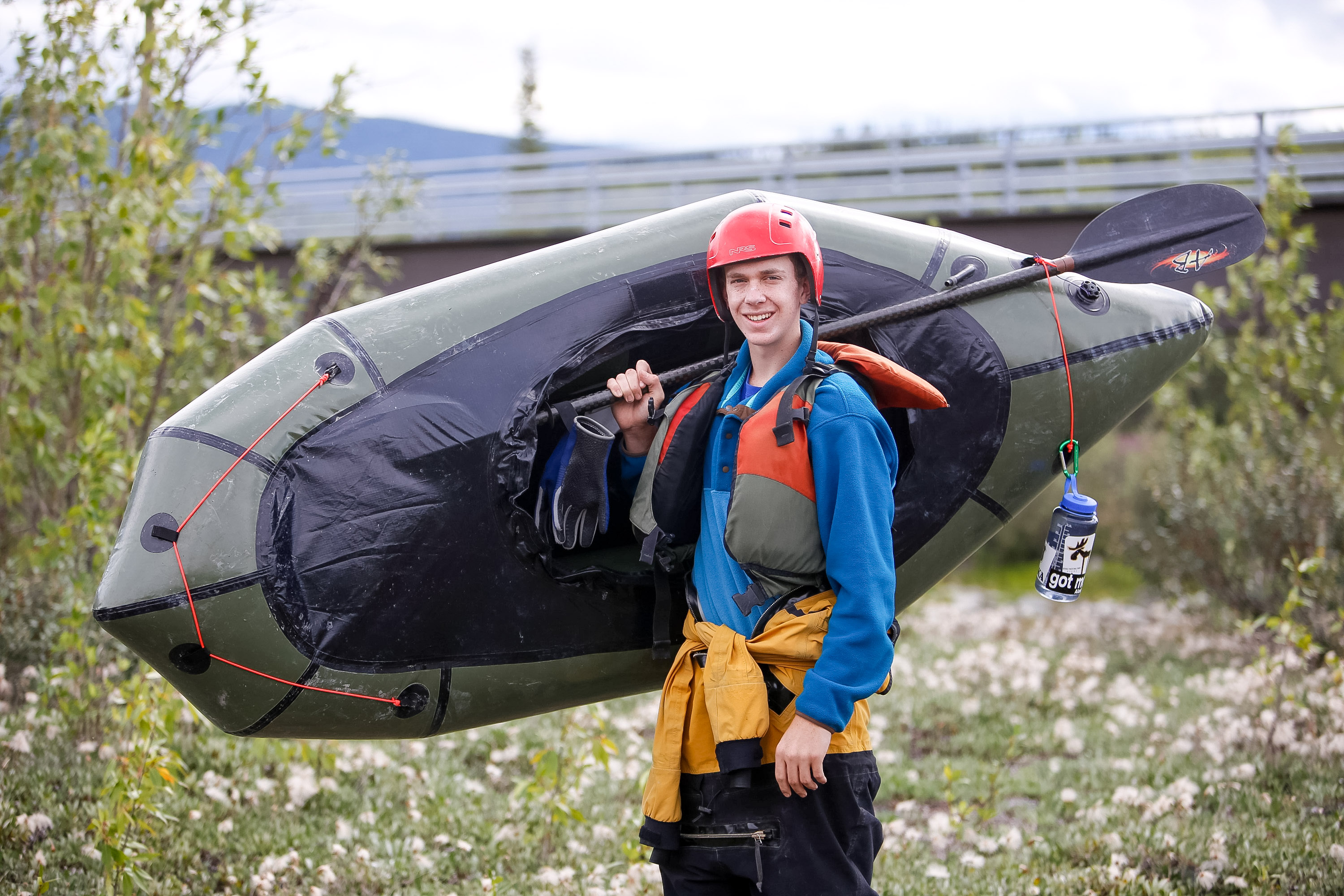 A student wearing a helmet carries a packraft and paddle across a grassy field