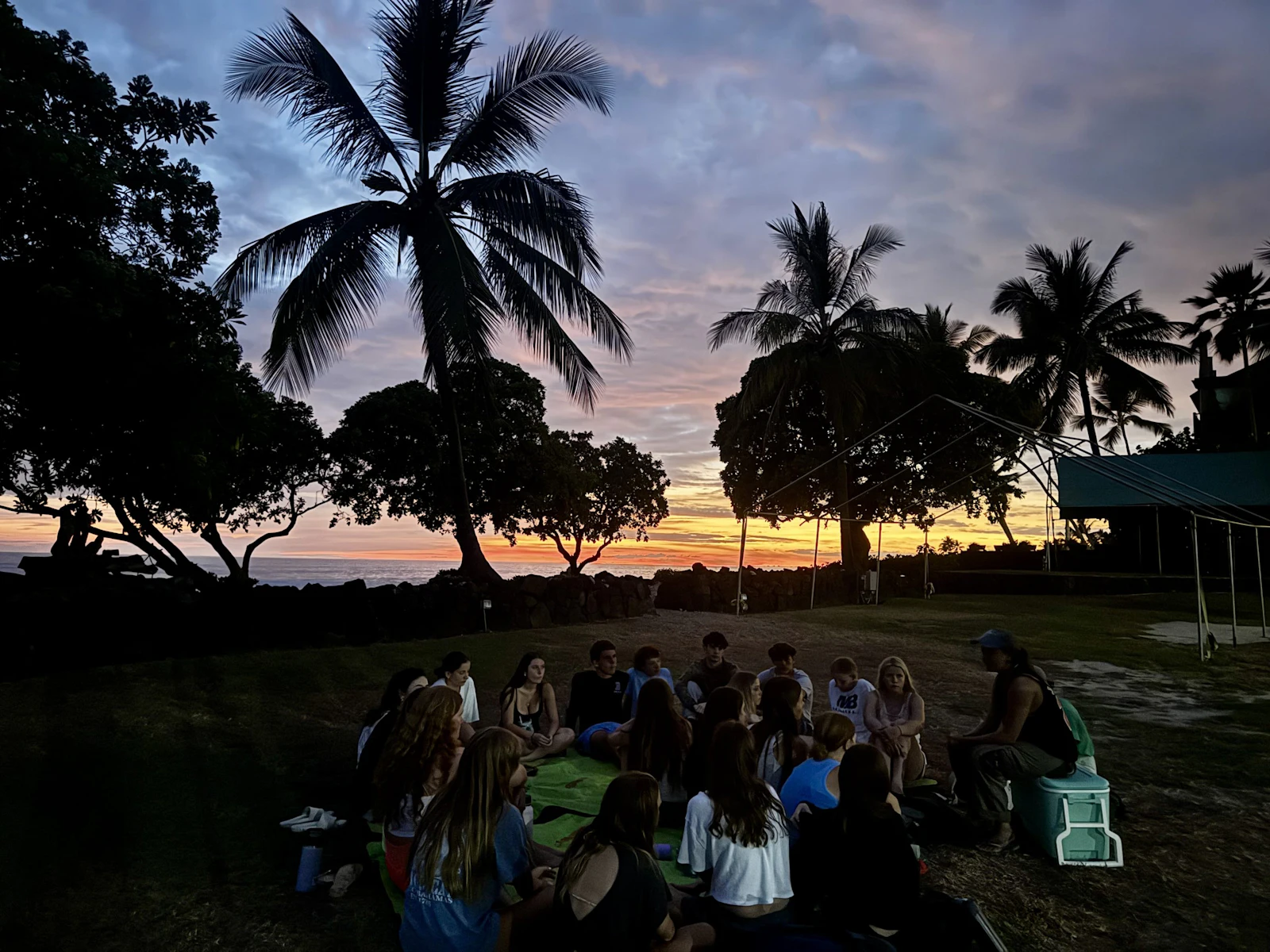 A large group of teen travelers sits in a circle on grass at sunset, silhouetted under coconut palms