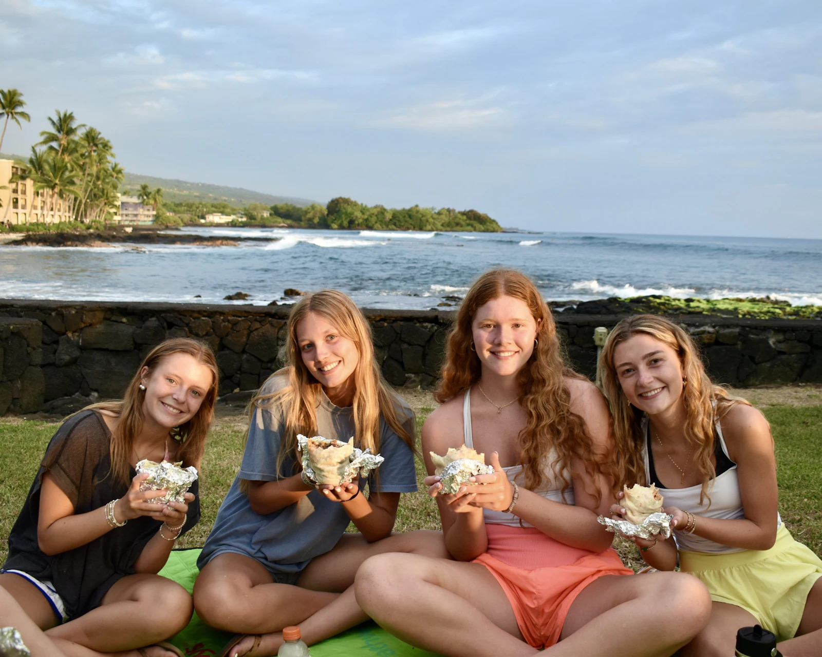 Four teen travelers sit on a blanket holding foil-wrapped burritos, a lava rock seawall and the ocean behind them
