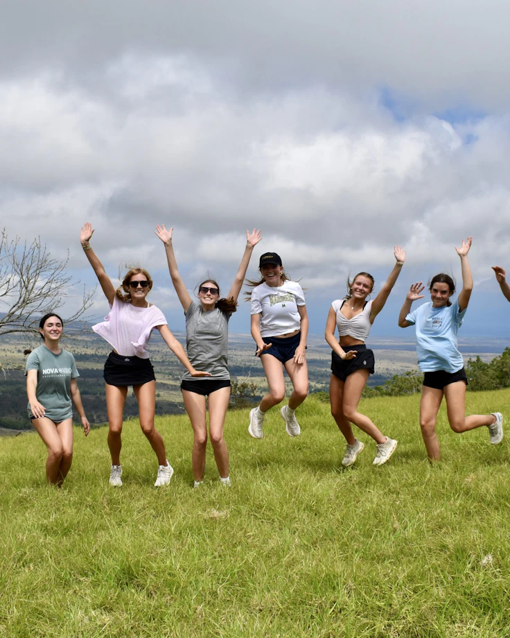 Nine teen travelers jump with arms raised on a green upcountry hillside in Hawaii under a bank of cloud