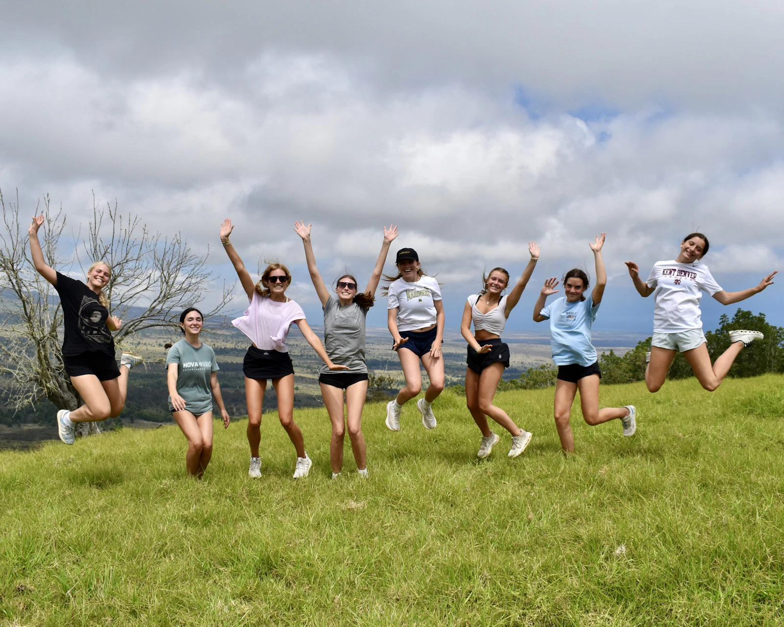 Nine teen travelers jump with arms raised on a green upcountry hillside under a bank of cloud