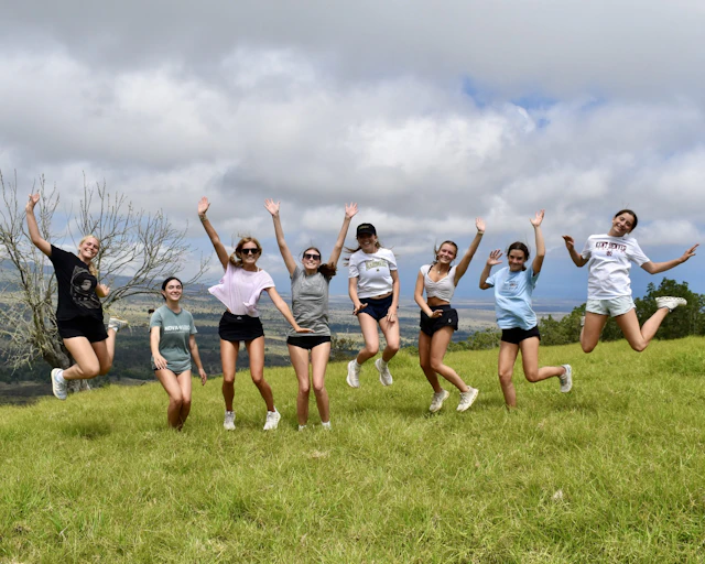 Nine teen travelers jump with arms raised on a green upcountry hillside in Hawaii under a bank of cloud