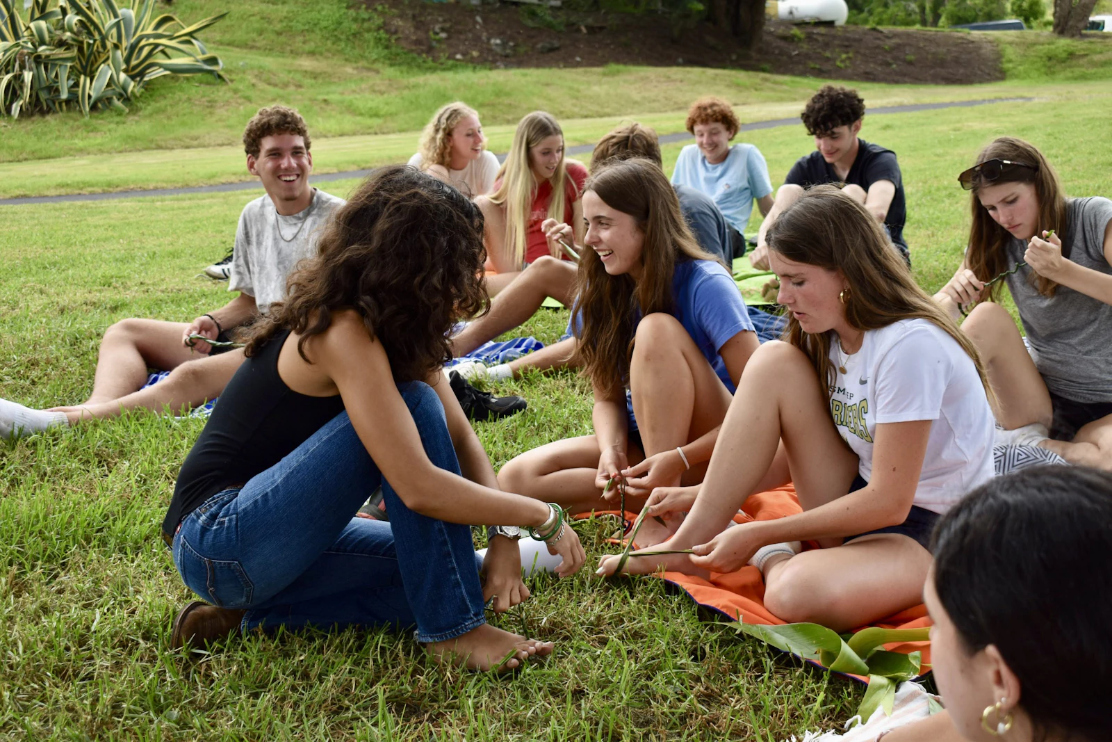 Teen travelers sit on the grass in a loose circle weaving long green leaves