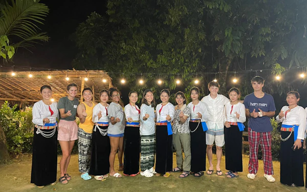 A long line of young people stand shoulder to shoulder on a covered wooden deck strung with festoon lights at night, several holding small gifts, with foliage behind them.