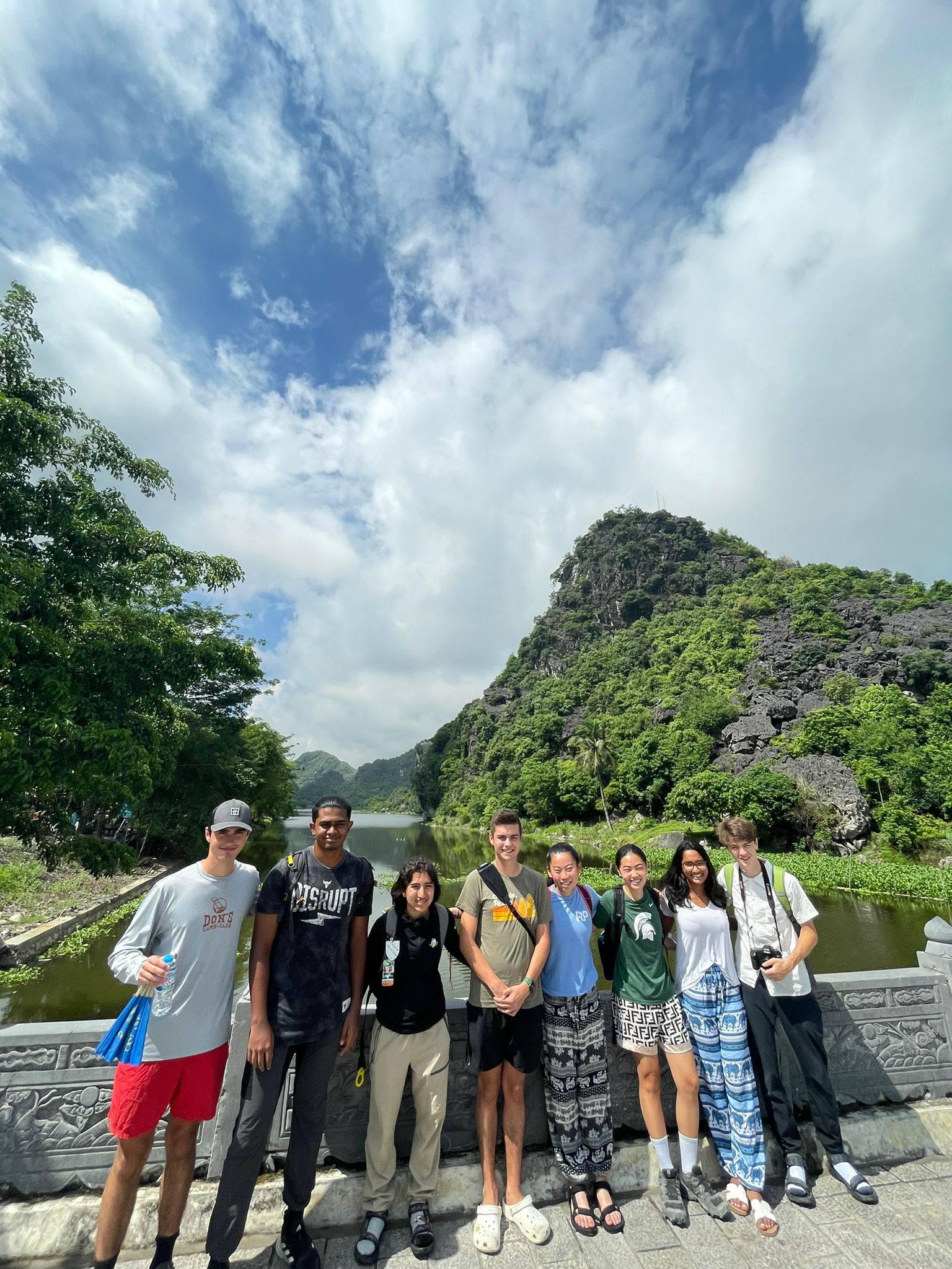 A group of young people stand together beside still water at the foot of a steep limestone peak covered in dense green vegetation, under a sky of tall white cloud.