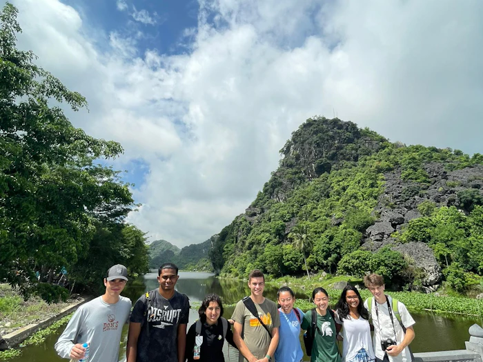 A group of young people stand together beside still water at the foot of a steep limestone peak covered in dense green vegetation, under a sky of tall white cloud.