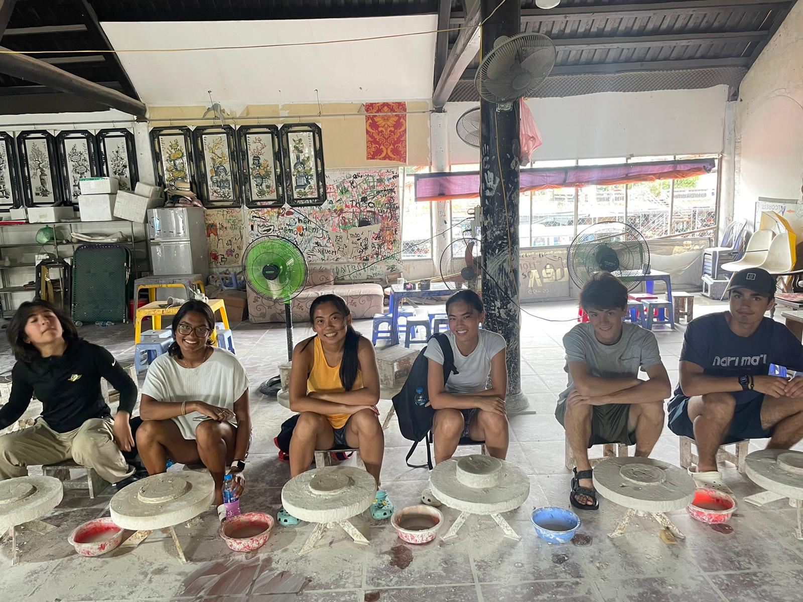 Young people sit on low stools in a row inside an open-fronted workshop, each working at a shallow basin set into a stone block.