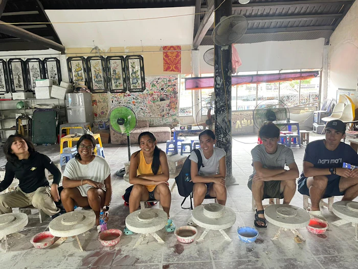 Young people sit on low stools in a row inside an open-fronted workshop, each working at a shallow basin set into a stone block.