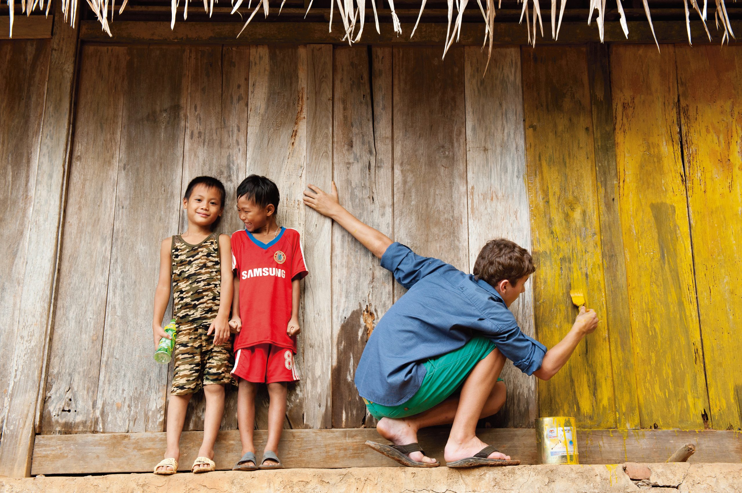 Volunteer painting a wooden wall while two local children watch in a Vietnamese village.
