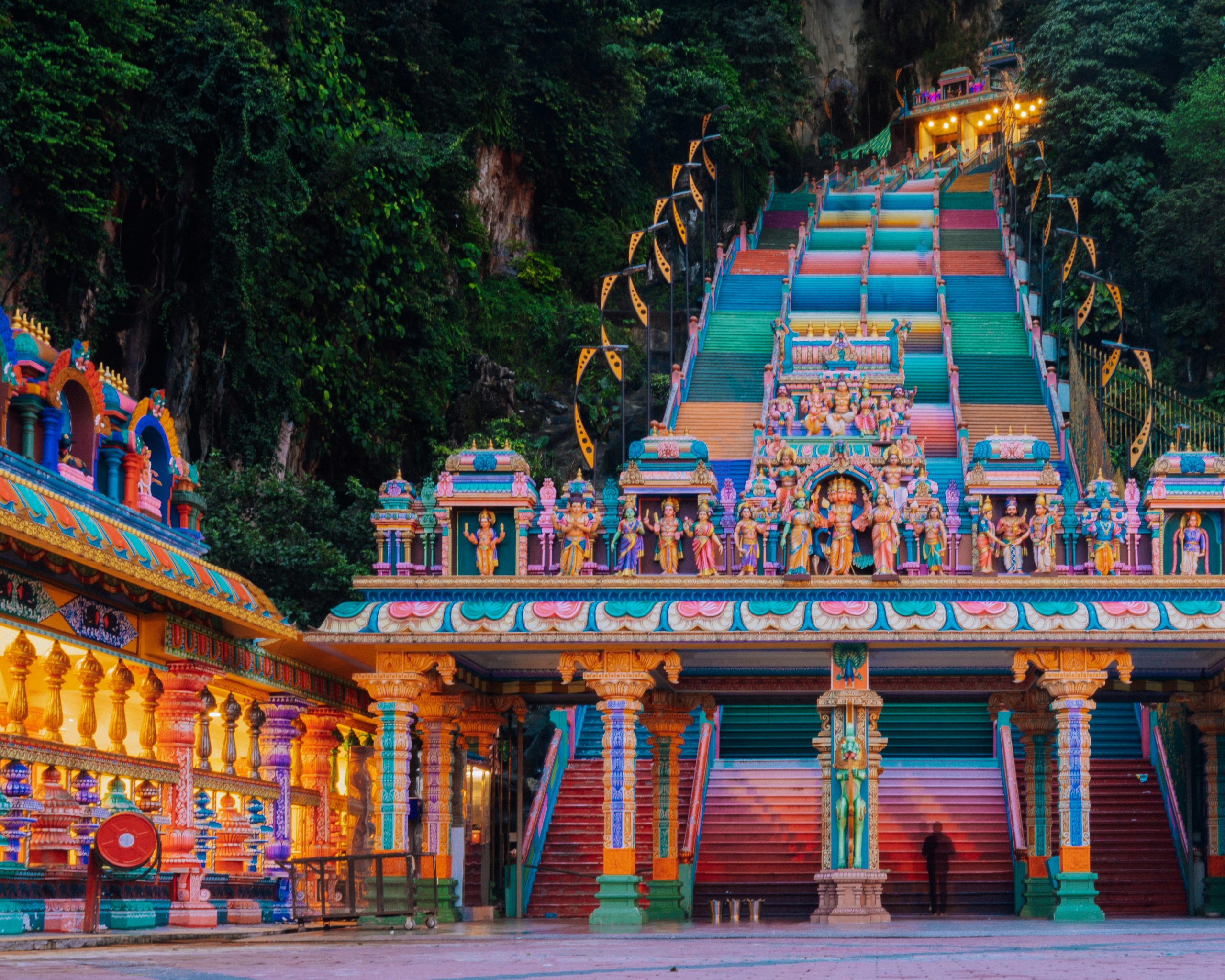 Colorful rainbow-painted stairs lead up to the ornate entrance of Batu Caves temple at dusk.