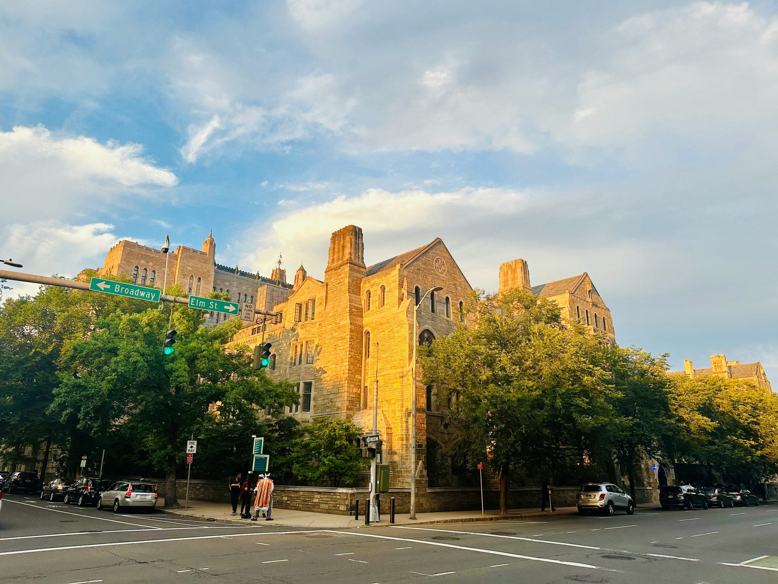 Gothic-style university buildings rise above a tree-lined street corner in the United States
