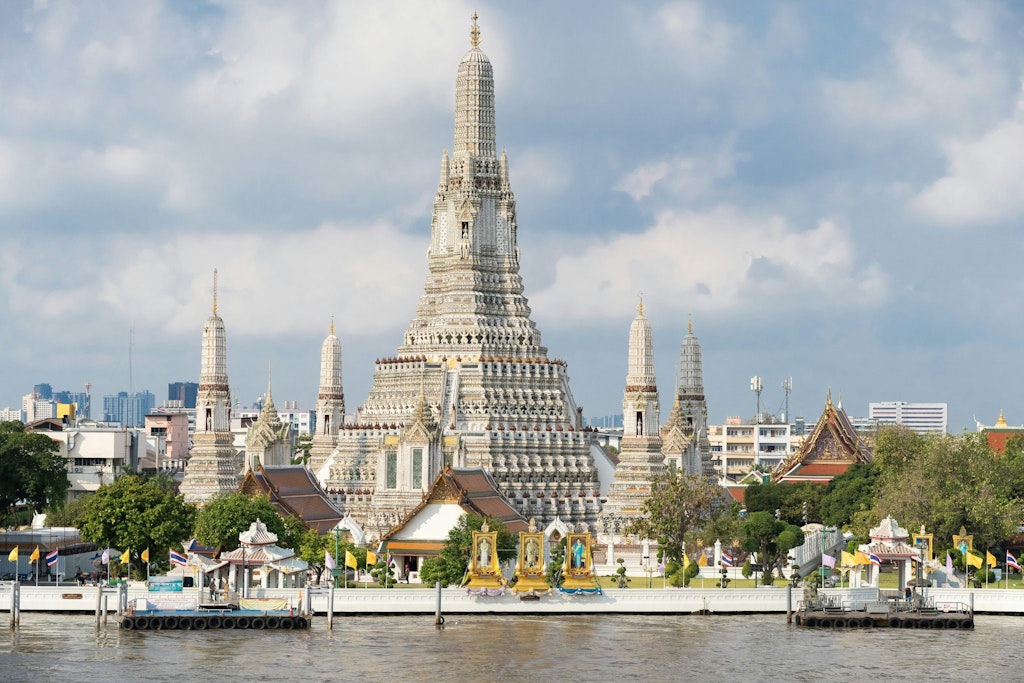 A image of Wat Arun (Temple of Dawn) in Bangkok, Thailand, features its tall spire adorned with intricate porcelain. The surrounded parks, houses and Chao Phraya River frame the temple against a cloudy and blue sky.