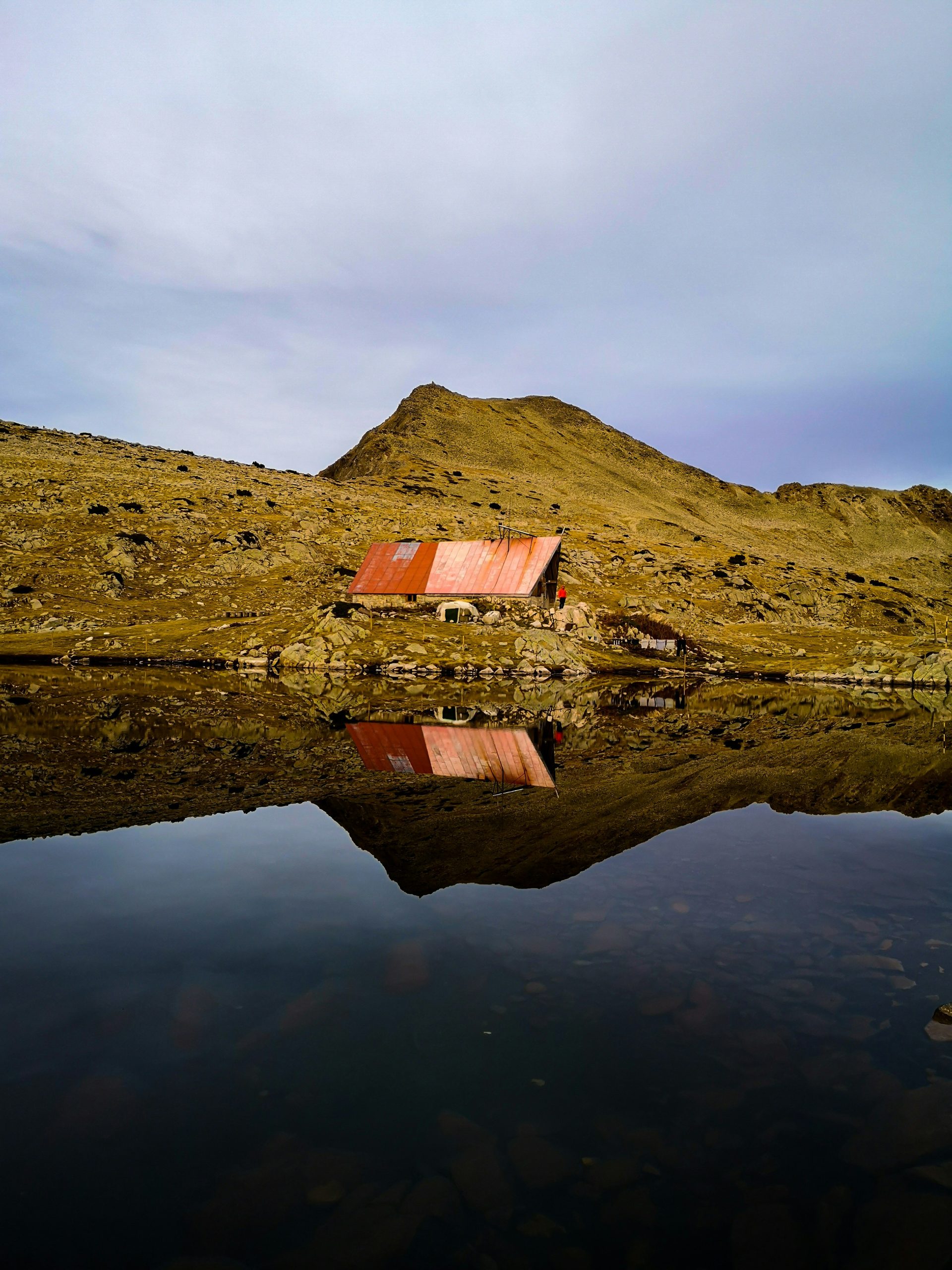 A hut with a rust-red roof beside a still lake below a bare summit in the Pirin Mountains.