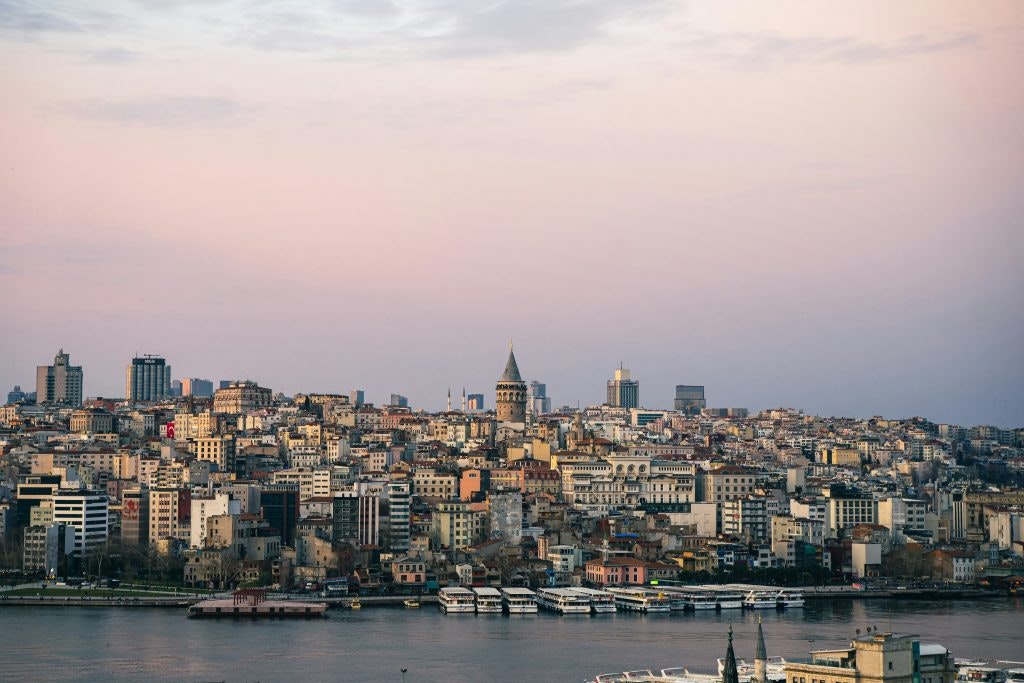 This image shows a broad view of the Istanbul skyline, dominated by the historic Galata Tower at the center. There is also a mix of residential and commercial buildings, with the waters of the Bosphorus in the foreground.