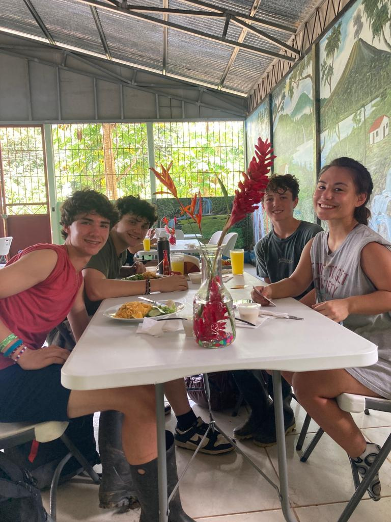 4 students (3 guys and 1 girl) seen sitting at a table, eating something which looks like breakfast or lunch. Inside a room in summer clothes and the sun coming through the windows showing the green nature waiting outside. All smiling for the photo